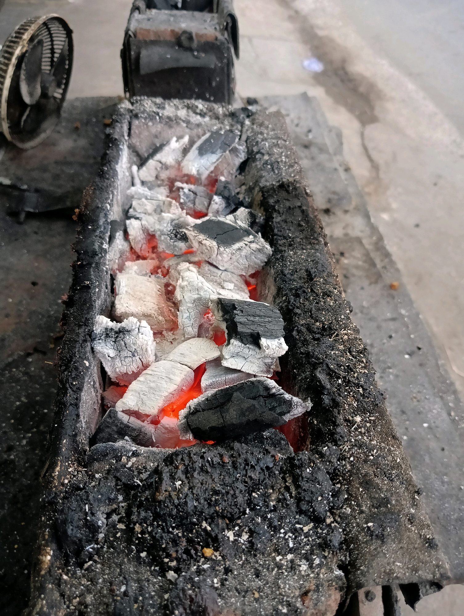 The image shows a close-up view of glowing charcoal in a rectangular brazier. The charcoal is predominantly white with some red embers visible underneath, indicating heat. The brazier itself appears to be made of metal with a dark, charred exterior covered in ash and debris. To the left of the brazier, partially visible, is a small, circular fan with a metal grille. The background consists of a grey, textured surface that appears to be concrete or pavement. The overall scene suggests an outdoor setting where the brazier is being used for cooking or heating. The location is identified as Sokaraja, Indonesia.