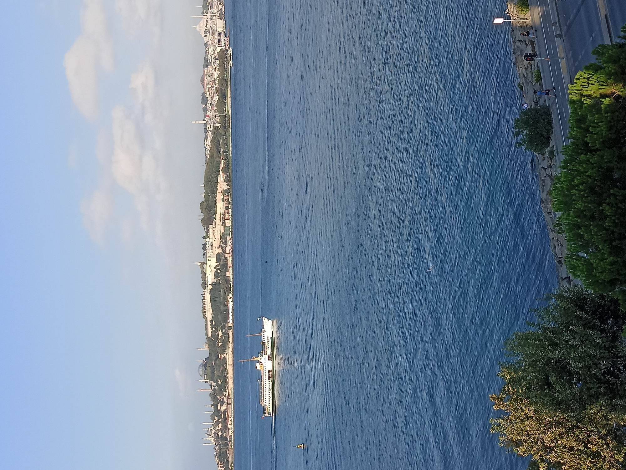 The image captures a panoramic view of the Bosphorus Strait in Üsküdar, Turkey. A white ferryboat, adorned with several decks and a flag at its stern, sails across the deep blue water from left to right. In the distance, the historic skyline of Istanbul is visible, featuring numerous minarets and domes of significant landmarks, including what appears to be Hagia Sophia. A small yellow buoy marks a navigational point in the foreground, to the left of the ferry. The water's surface is rippled, suggesting a gentle breeze. The foreground of the image is dominated by lush green trees and foliage, with a rocky embankment leading down to the water's edge. Further along the embankment, a road is visible with a few individuals walking and a traffic light. The sky is a clear blue with scattered white clouds.