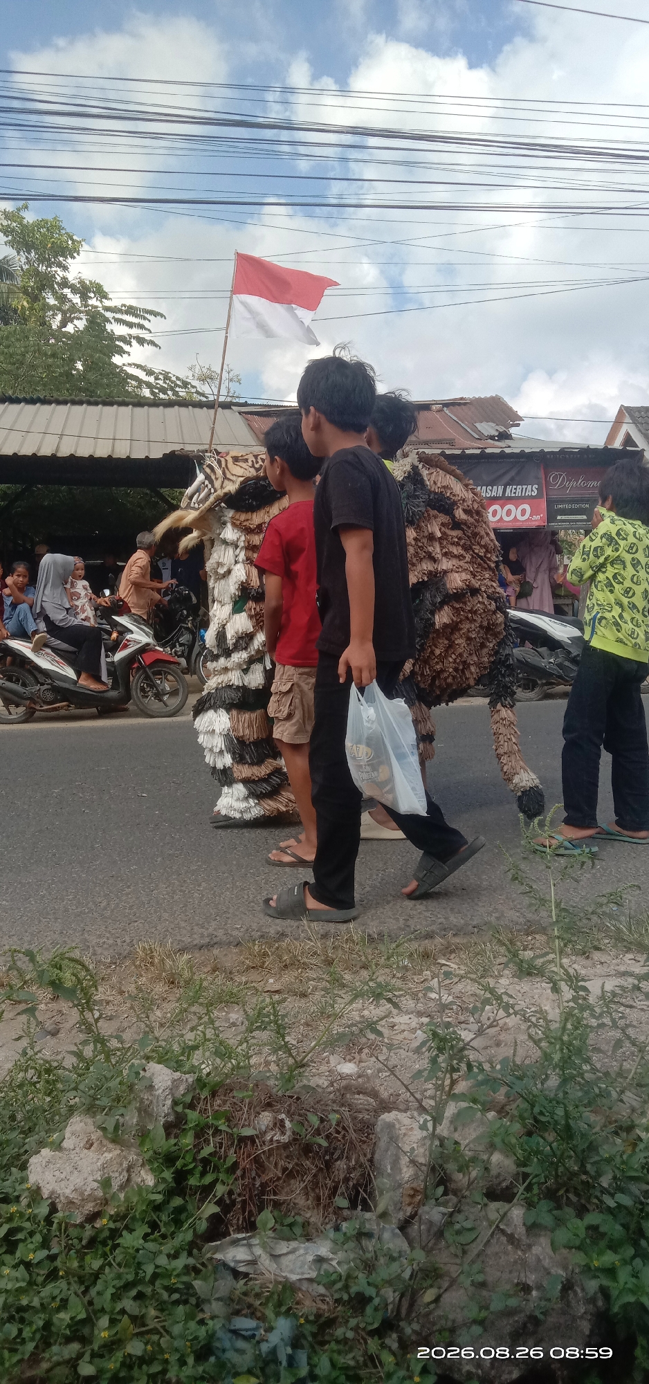 In Pakong, Indonesia, a group of people are walking down a street, participating in what appears to be a parade or cultural event. At the forefront, a person in a elaborate costume resembling a tiger is being led. The costume is made of what looks like straw or fabric, with black, white, and brown stripes and a furry mane. Following the costumed figure are several children, with one boy in a black t-shirt holding a transparent plastic bag. Behind them, a person in a yellow hoodie with a distinctive pattern is walking. In the background, a red and white Indonesian flag waves from a pole. Other individuals are visible on the side of the road, with one woman wearing a hijab seated on a motorcycle and a man standing behind her. A banner with text, including the word "DIPLOMA" and "000.000", is displayed on a building behind the participants. The scene takes place on a paved road, with grassy areas and vegetation bordering the street. The sky above is partly cloudy. The timestamp "2026.08.26 08:59" is visible in the lower right corner of the image.