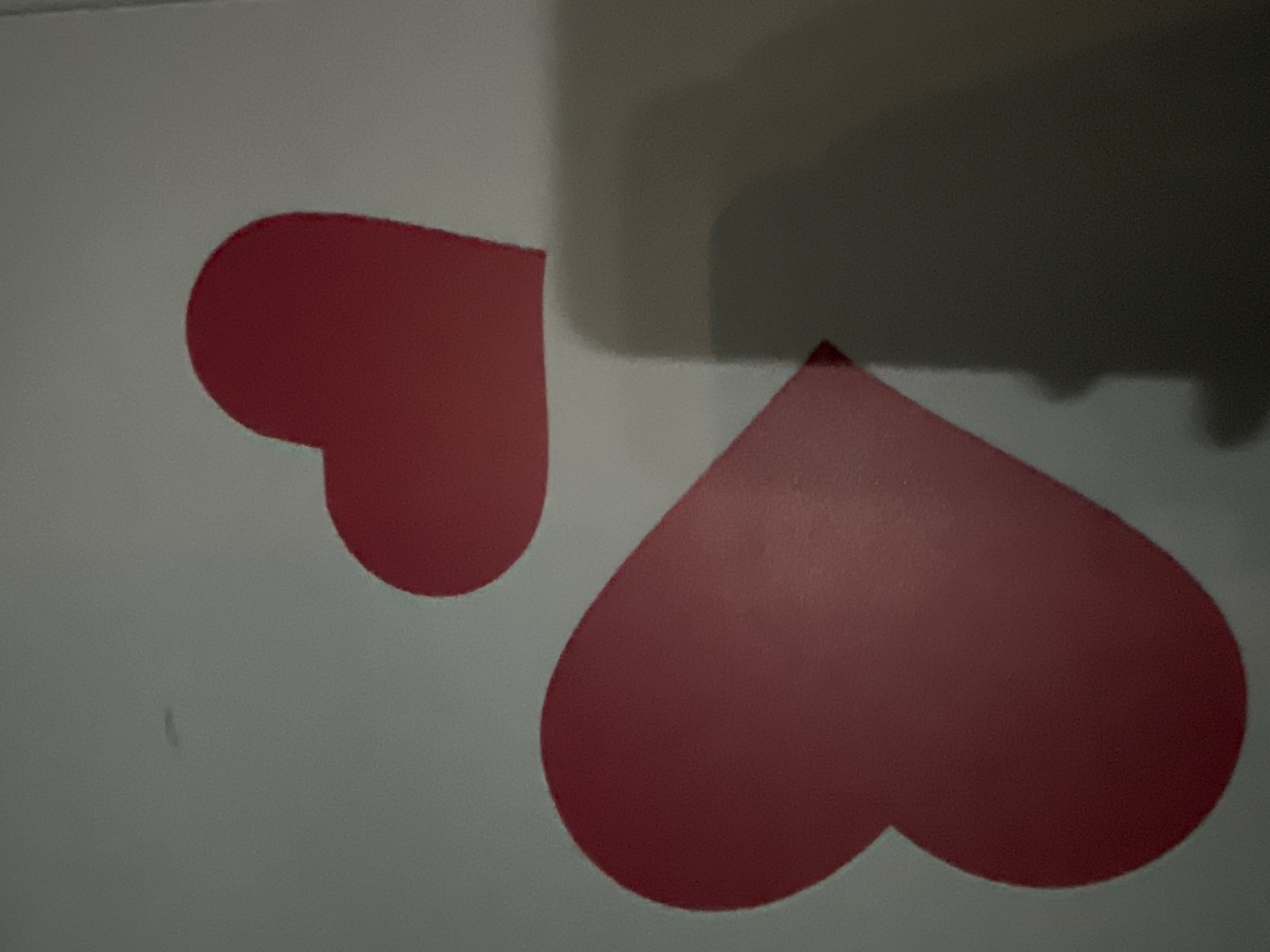 The image displays two red heart-shaped stickers on a white surface. The larger heart is positioned on the left side of the frame, while a smaller heart is located to its right. A shadow from the larger heart is cast on the surface below it. There are no people or other objects visible in the image. The setting appears to be indoors, with the white surface likely being a wall or a large flat object. The lighting suggests a single light source, casting a distinct shadow. The background is a plain, light gray. The image does not provide any information about the city of Yuzhong District, China.
