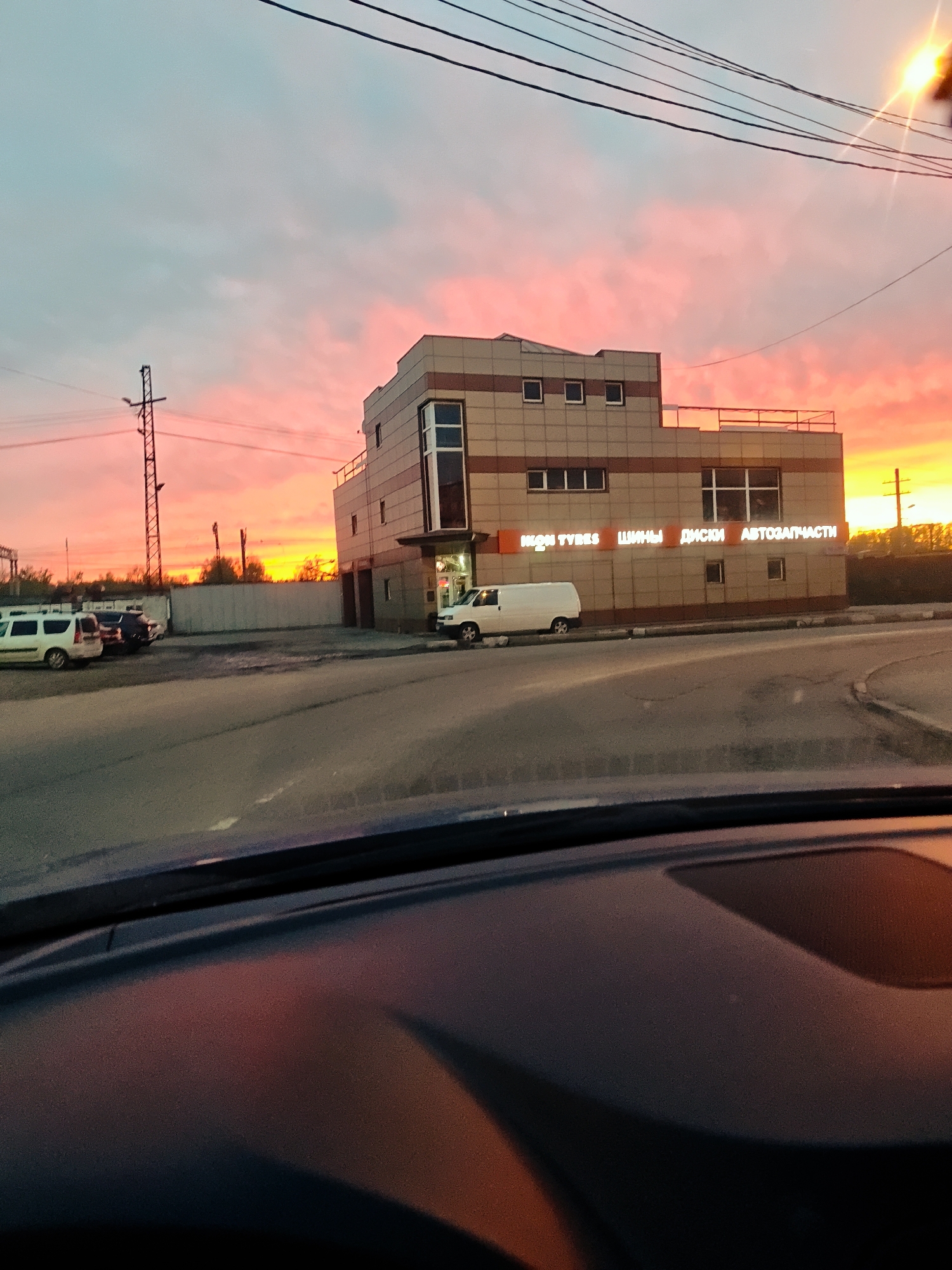 View from inside a vehicle looking towards a two-story building in Dmitrov, Russia. The building has a beige facade with brown accents and signage in Russian that reads "IKON TYRES," "SHINY," "DISKI," and "AVTOZAPCHASTI" (Tires, Wheels, Discs, and Auto Parts). A white van is parked in front of the building, and other cars are parked in the lot to the left. A utility pole stands to the left of the building. The sky is brightly colored with hues of pink, orange, and yellow, indicating sunrise or sunset. A portion of the vehicle dashboard is visible at the bottom of the image.
