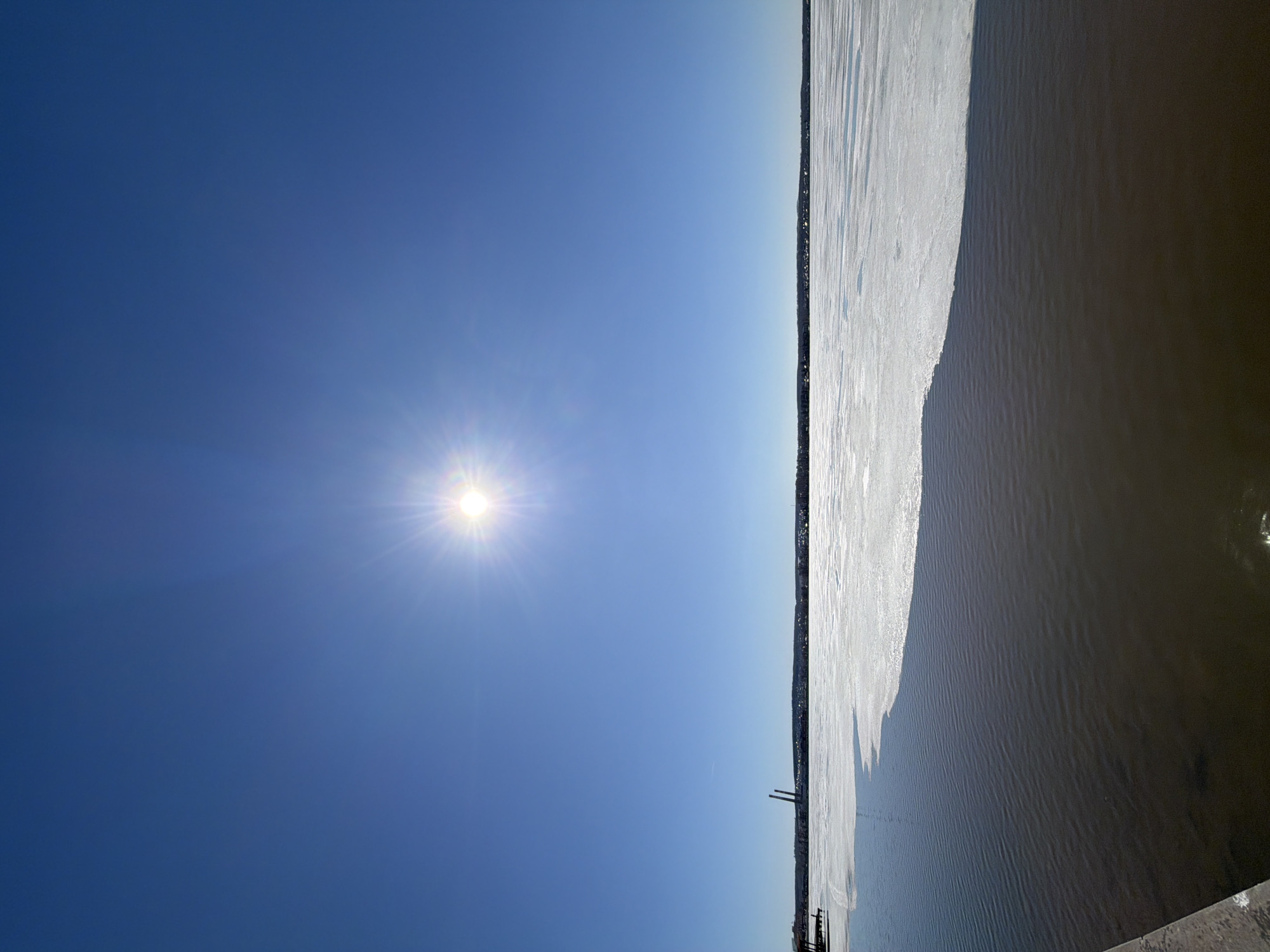 The image depicts a clear, sunny day at a river in Votkinsk, Russia. The sky is bright blue with the sun shining intensely. On the right, the river is partially frozen, with ice covering part of its surface. The other part of the river appears as open water. A thin line of trees is visible along the edge of the ice. In the distance, there are two tall smokestacks near what is likely a pier. No people are visible.