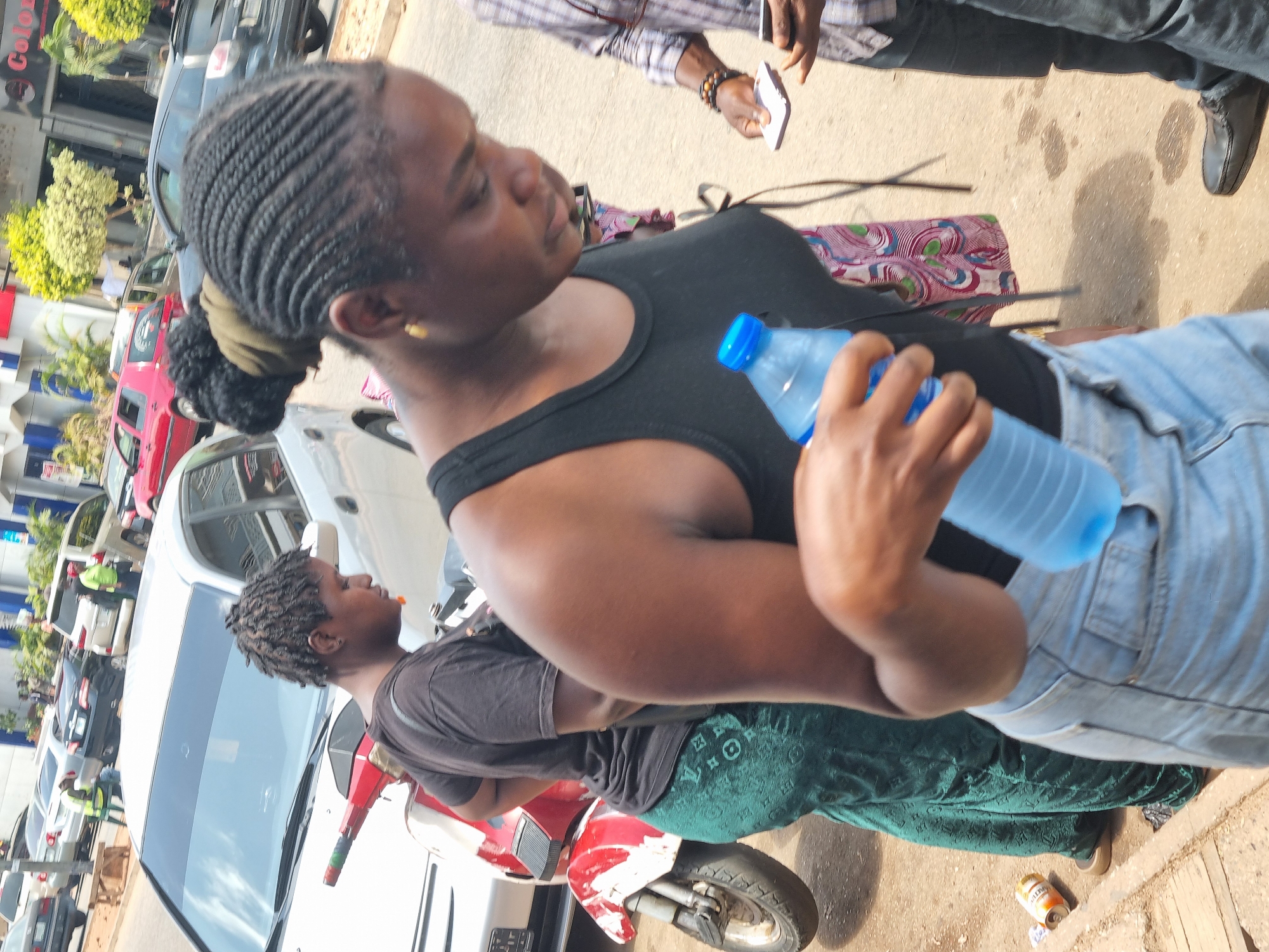 The image shows two Black women in an outdoor setting in Jos, Nigeria. The woman in the foreground is holding a blue water bottle. She has braided hair pulled back into a bun, and is wearing a black tank top and blue jeans. Another woman is seated behind her on a red and white scooter. She is wearing a black t-shirt and a long green skirt with a patterned design. The scooter is parked next to a white car. The background includes several other vehicles, a building with storefronts, and some greenery. The ground appears to be paved. There is a discarded drink can near the scooter. A man in a plaid shirt is partially visible in the upper right corner of the frame.