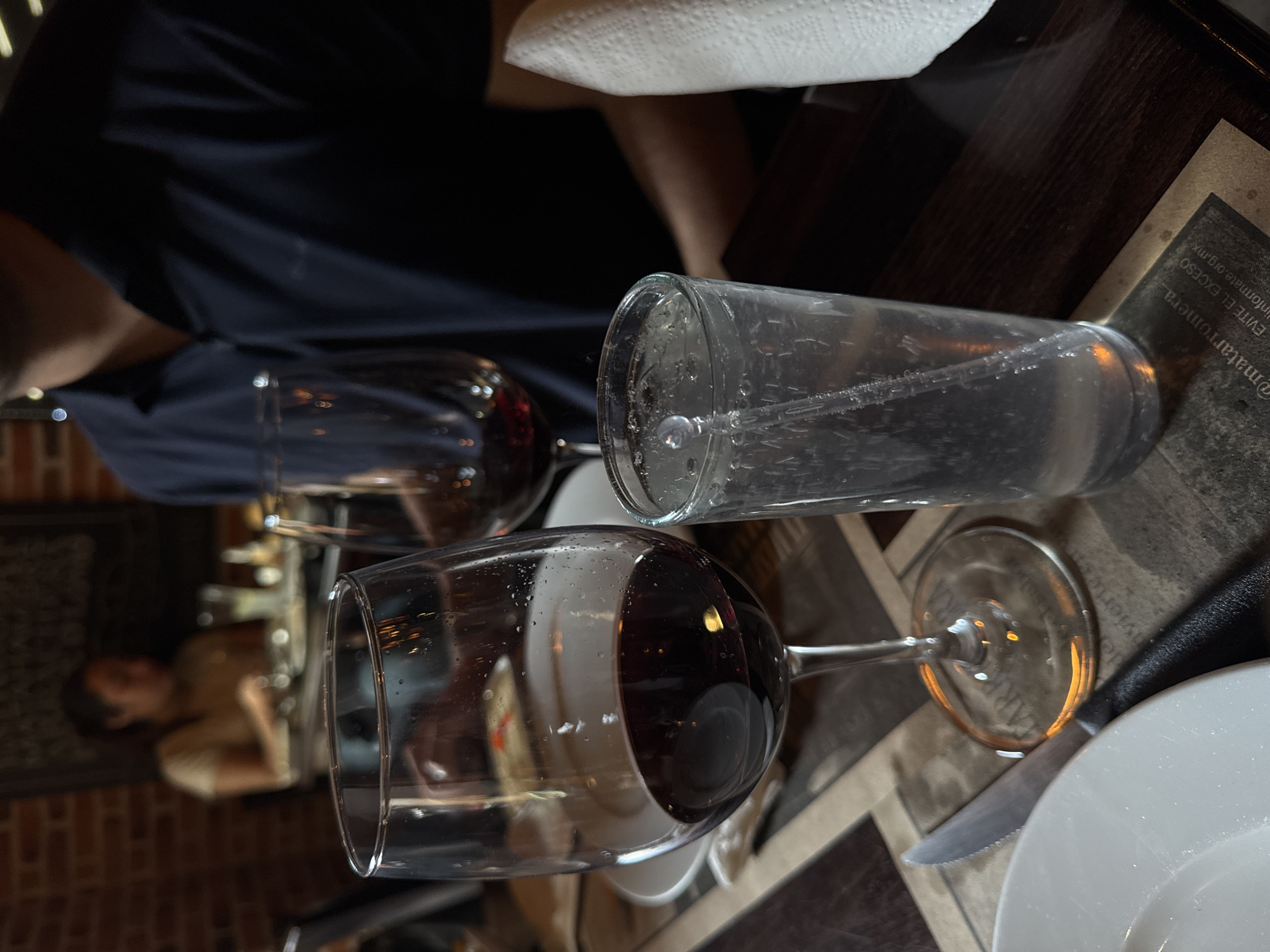 A table in a restaurant in Puebla, Mexico, is set with a white plate, a knife, a glass of water with a straw, and two wine glasses, one filled with red wine. A person wearing a dark blue shirt is seated at the table, and another person is visible in the background. The table has a dark, possibly wooden, surface with a printed placemat. The overall lighting is dim.