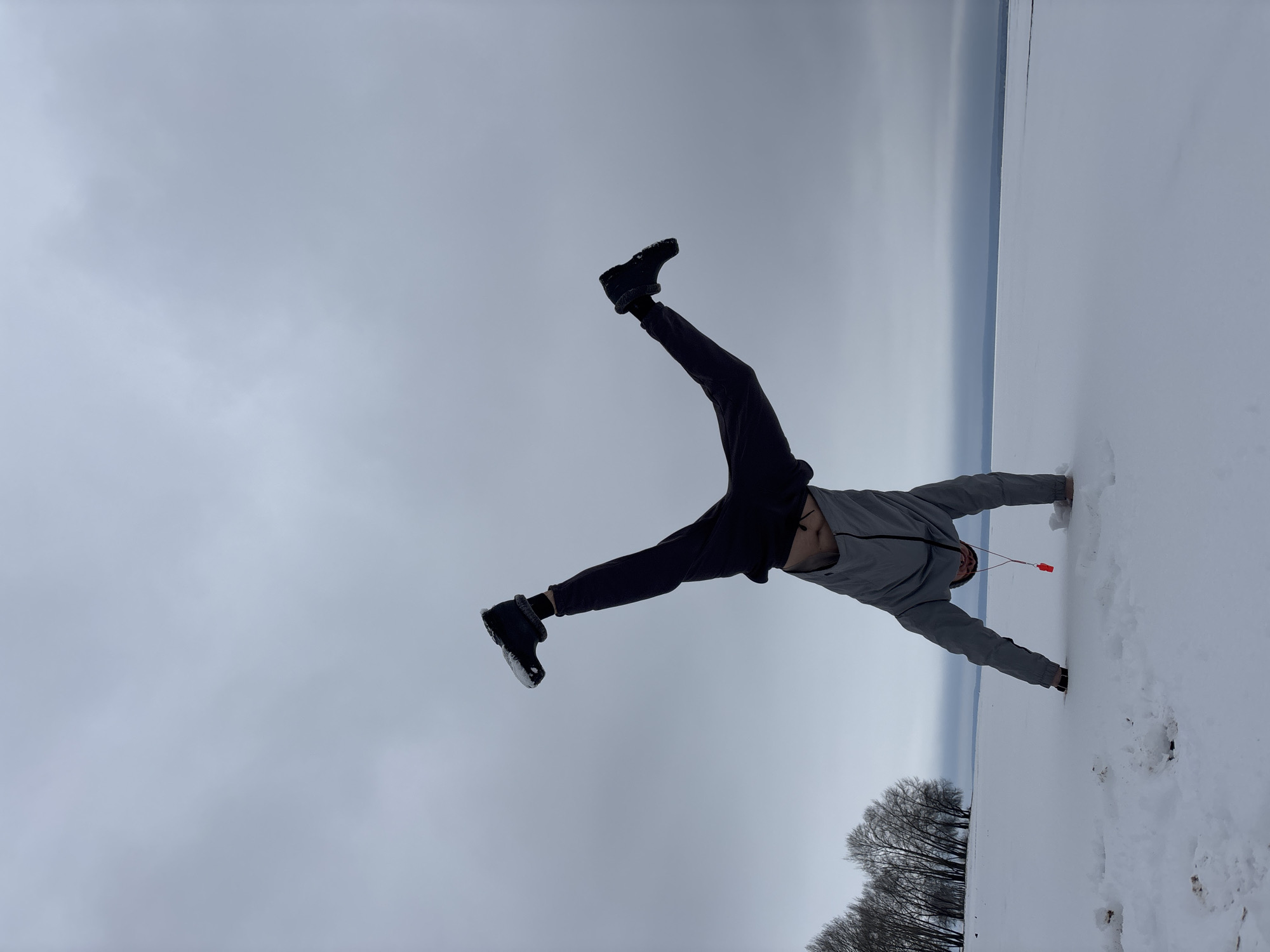 A person in dark pants and a grey jacket is doing a handstand in the snow. The location appears to be a snowy field with trees visible in the background. The sky is overcast and there is what appears to be a body of water or open field in the background. There's no definitive indication of the city of Noviy, Russia, although the climate is consistent.