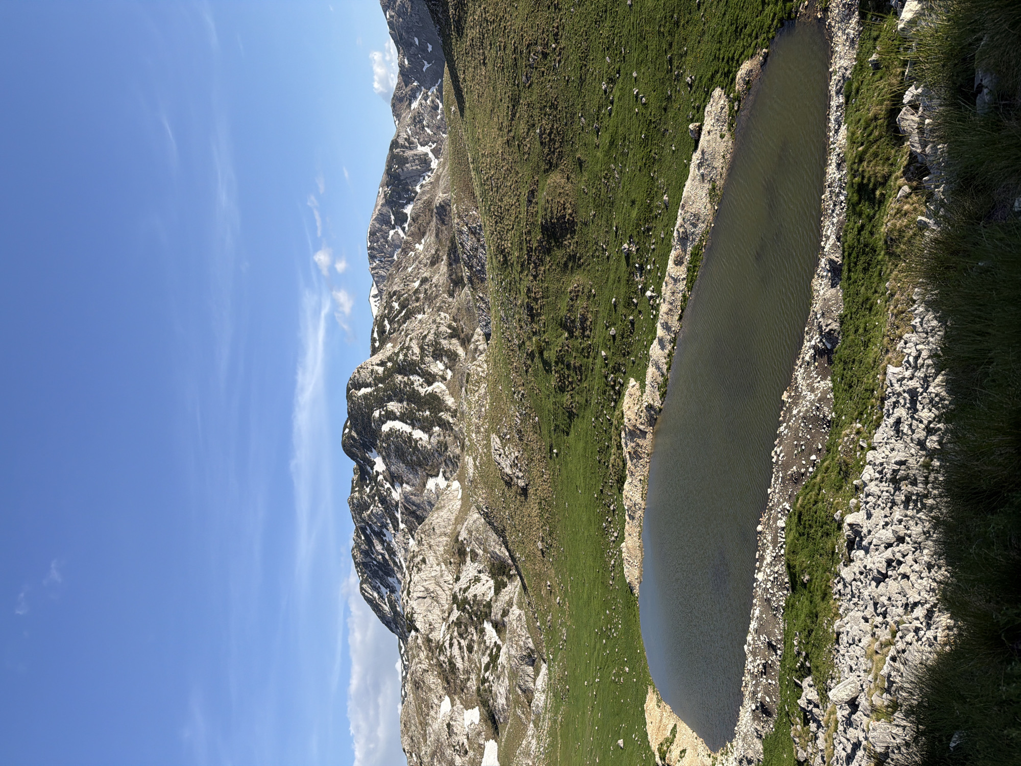The image depicts a mountainous landscape in Boričje, Montenegro. A long, dark lake is situated along a grassy hillside that ascends towards a rugged, rocky mountain. Patches of snow remain on the mountain's rocky slopes. The sky is blue with scattered wisps of clouds. The vegetation appears lush in areas, contrasting with the barren rocky terrain. No people are visible.