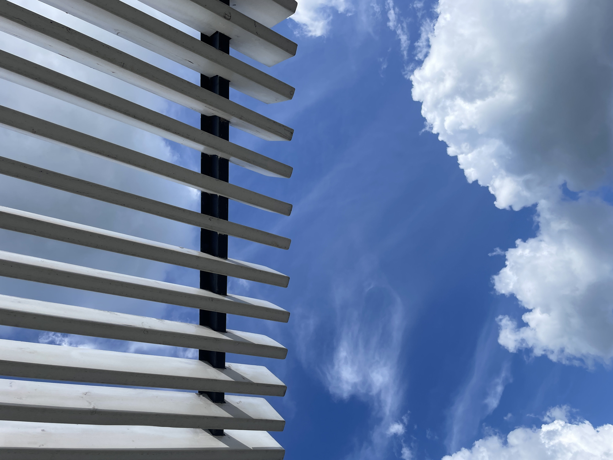 An angled upward shot captures a bright blue sky dotted with white clouds. White wooden slats, part of an overhead structure like a pergola or canopy, form a repeating pattern across the top of the frame. These slats are supported by a dark, unseen structure. The scene evokes a sense of being outdoors on a clear day, possibly in a park or residential area. The location is Laishevo, Russia.