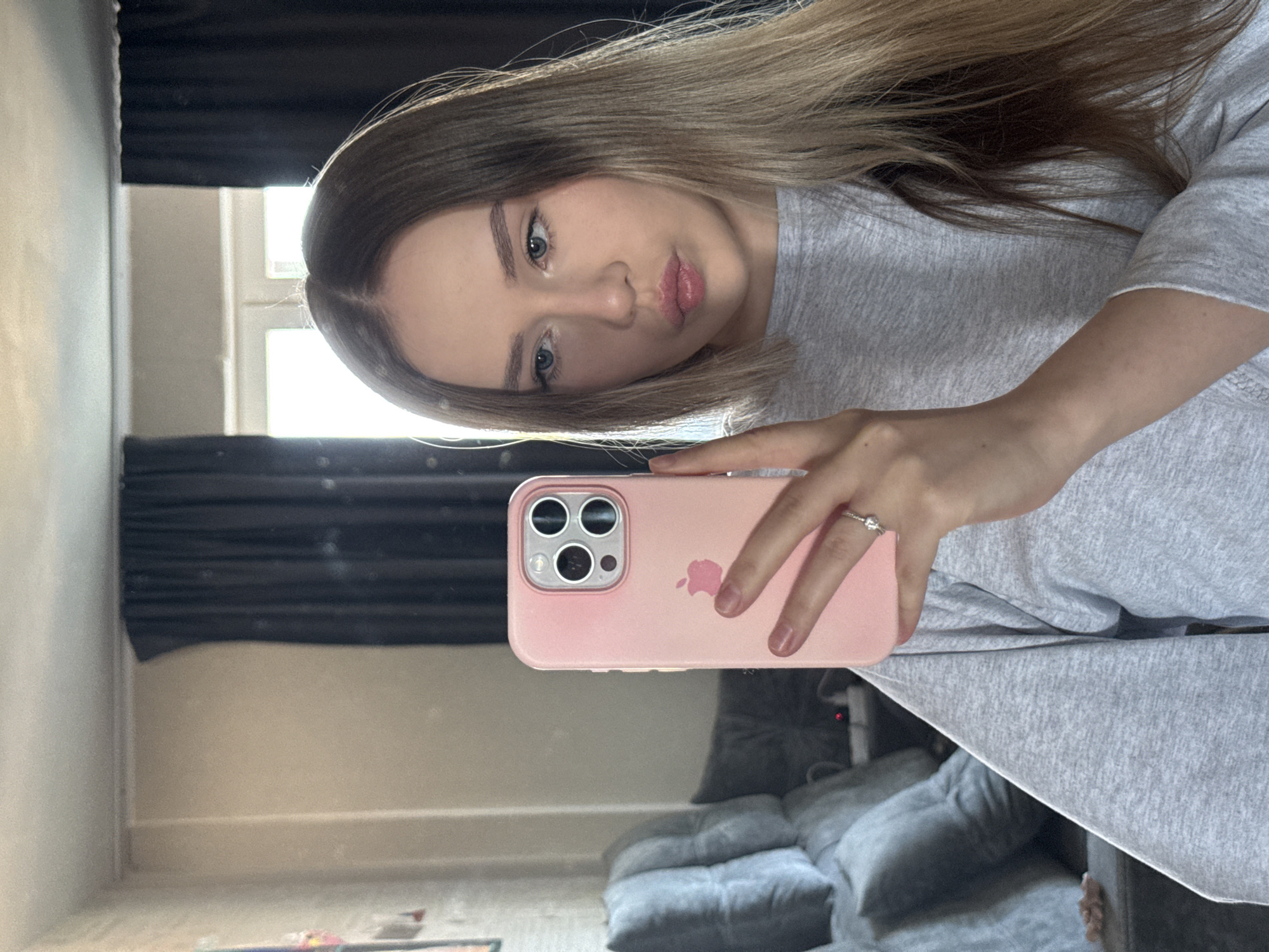 A young woman is taking a selfie in front of a mirror. She is fair-skinned, with light brown hair with blonde highlights. She is wearing a light gray sweatshirt. In her right hand, she holds a pink iPhone in a matching pink case. The reflection shows a living room with a gray couch and dark curtains. The exact location in the city of Chaikovsky, Russia, cannot be determined from the provided image.