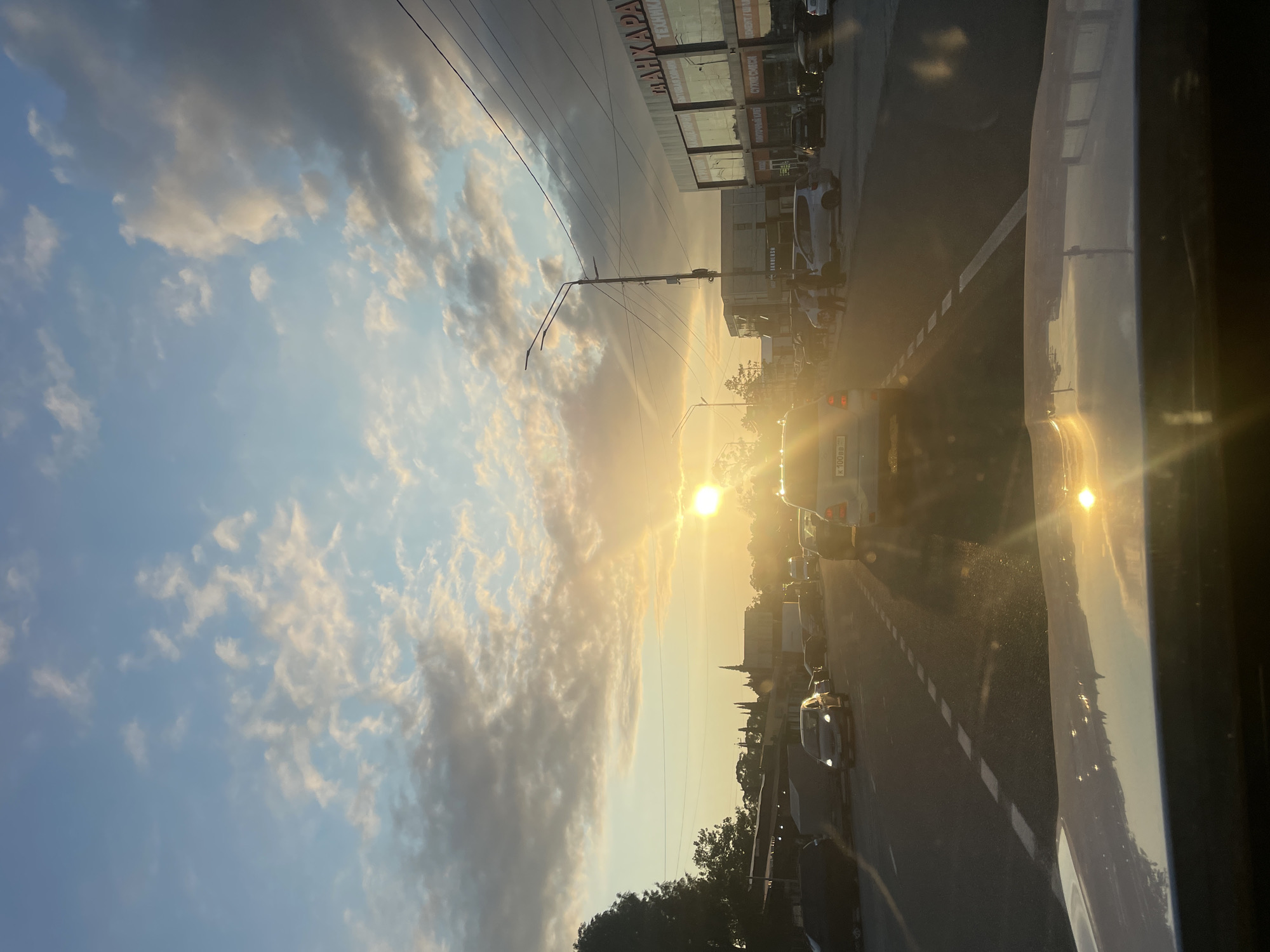 The image captures a street scene in Adzhiryuara, Abkhazia, during sunset. The sun is low on the horizon, casting a warm, golden light across the sky and the environment. Numerous clouds are present, ranging from bright white illuminated by the sun to darker gray ones. A street with multiple lanes and visible lane markings is depicted, with several cars driving on it. The foreground shows the hood of a vehicle, reflecting the sunset. On the right side of the street, there are buildings with signage, suggesting commercial establishments. Some trees are visible on the left side of the scene. The overall atmosphere is that of a typical evening commute or a quiet street at dusk.