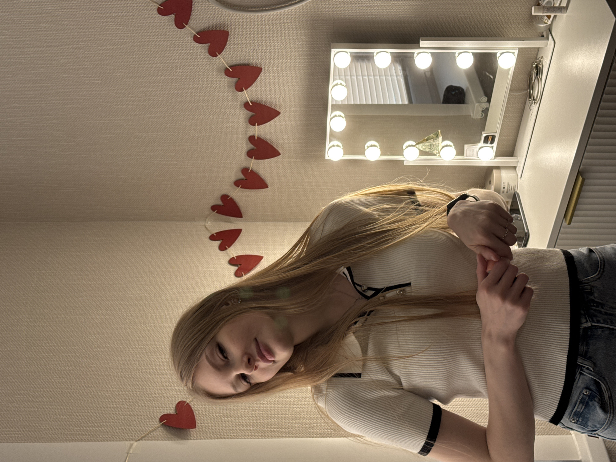 A young woman with long blonde hair is in a room with light beige walls. She is wearing a white, ribbed, short-sleeved shirt with black trim and blue jeans. A string of red heart-shaped decorations is visible near the ceiling. There is a white vanity with a lighted mirror attached to the wall. The location is possibly in Perm, Russia, given the prompt.