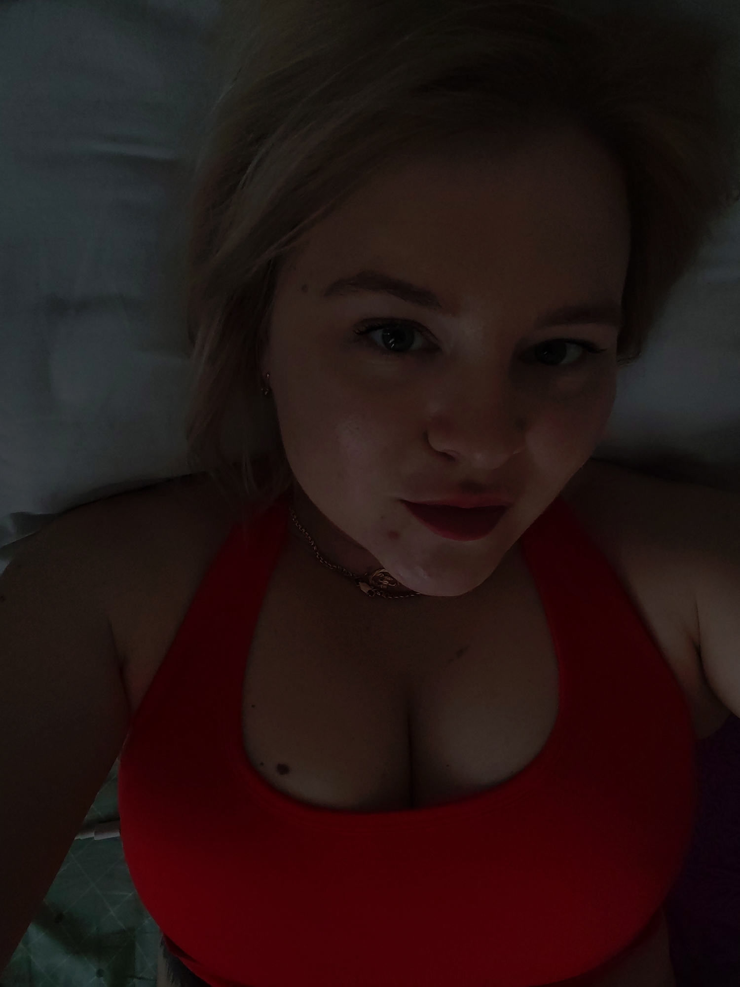 A blonde woman with fair skin and red lipstick is seen from a high-angle perspective. She is wearing a red halter top. She is indoors. There is a mole visible on her chest. A gold chain necklace adorns her neck. The background appears to be a bed. The location is unknown.