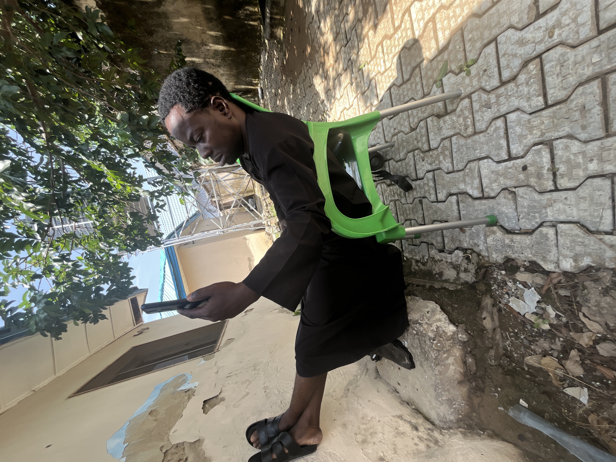 A person is seated on a green plastic chair outdoors in what appears to be Galadimawa Village, Nigeria. The individual is wearing a black robe and black sandals, and is holding a mobile phone. Their legs are crossed, with one foot resting on a textured, light-colored wall. The ground is paved with interlocking concrete blocks, and there are fallen leaves and debris scattered around. Overhanging tree branches with green leaves create shadows on the ground. In the background, there is a building with a light-colored, peeling facade and a window, as well as other structures with blue roofs and metallic scaffolding. The lighting suggests it is daytime, with sunlight casting distinct shadows.