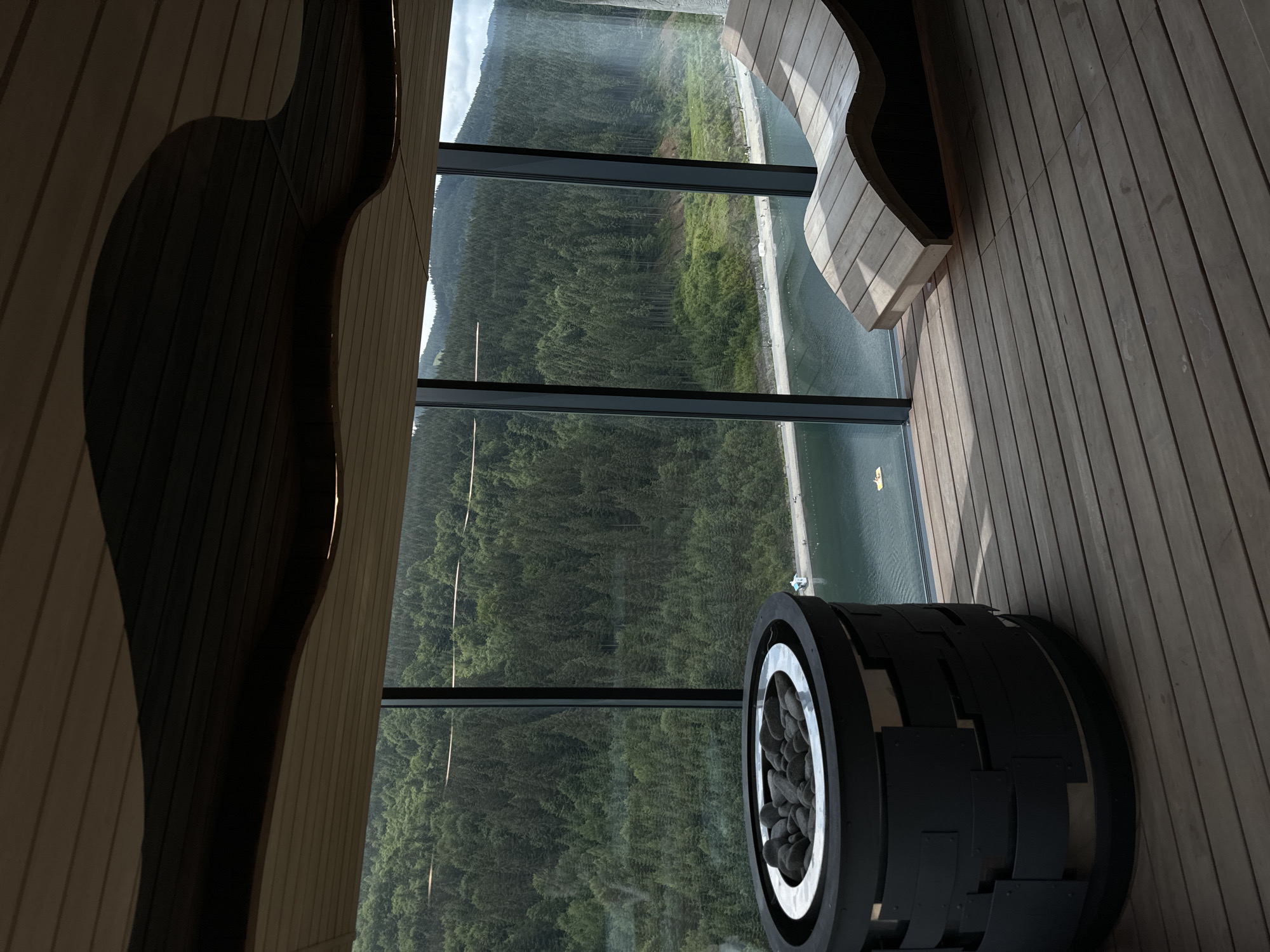 An interior view from what appears to be a modern spa or sauna facility in Polianytsia, Ukraine, showcases a large, floor-to-ceiling window offering a panoramic vista of a densely forested mountain range and a body of water. In the foreground, to the left, sits a dark, cylindrical object filled with dark stones, possibly a sauna stove or a decorative element. To the right, a curved wooden lounge chair is positioned on a wooden deck. The expansive window reveals a vast expanse of green trees covering rolling hills, with a calm body of water visible in the middle ground. Along the edge of the water, a sandy strip with several small figures suggests a beach or walkway. A single yellow inflatable object is visible on the water, and a small white structure can be seen on the shoreline. The ceiling is made of light-colored wooden planks with dark accents and lighting fixtures embedded within.