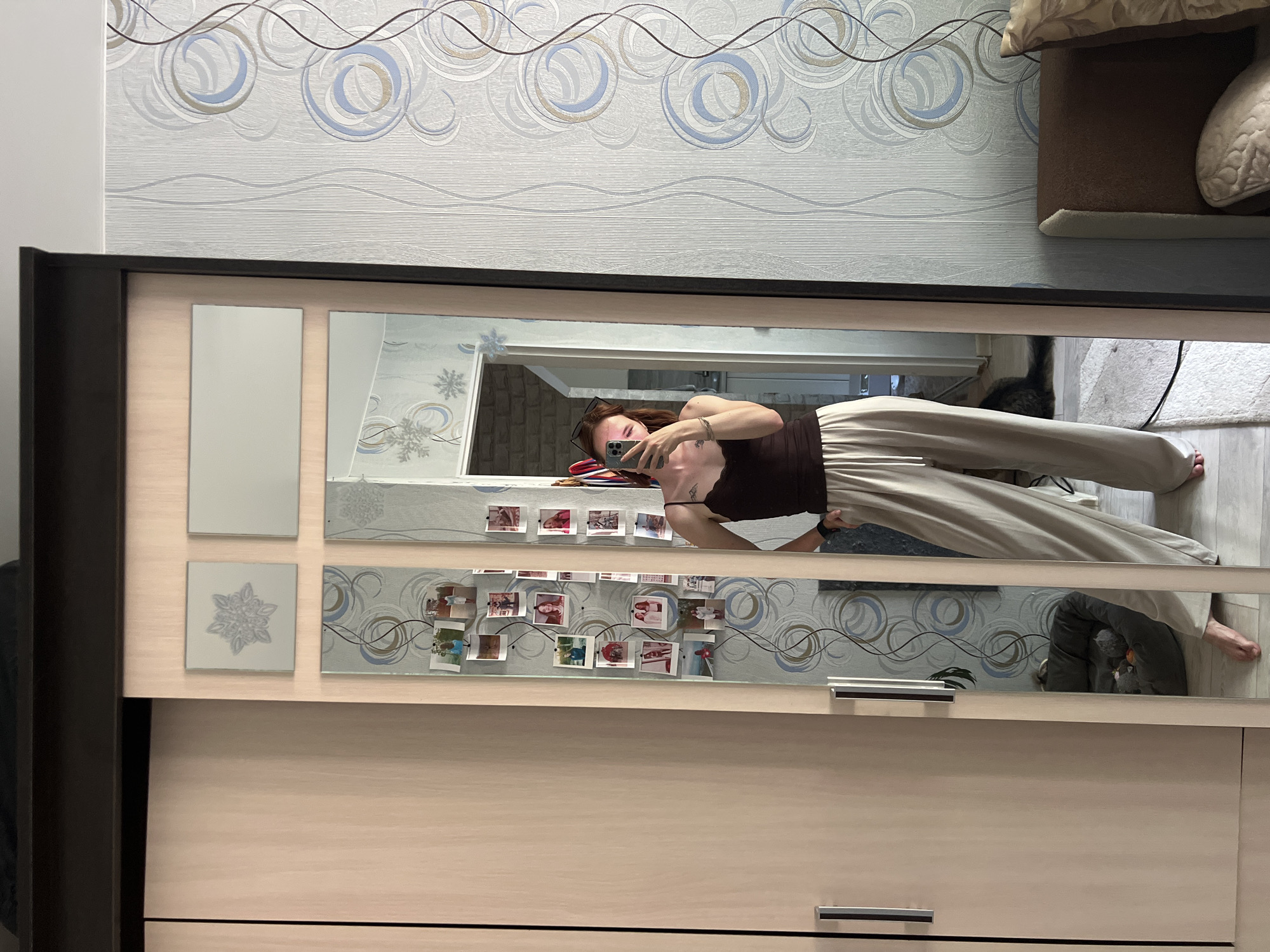 The image shows a full-length mirror reflecting a young woman in a dark tank top and light-colored wide-leg pants. She is holding a smartphone and taking a selfie. The mirror is part of a wardrobe with light wood doors. The wall behind the mirror is covered in light blue and white patterned wallpaper. A cat bed with a toy is visible in the reflection on the floor near the wardrobe. The room appears to be a bedroom or living area. The location of Chaikovsky, Russia is not discernible from the image itself; this is external information.