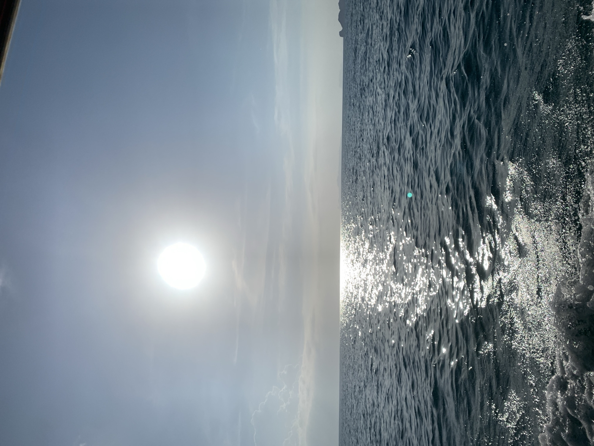 The image depicts a bright, sunny day over the ocean. The sun is high in the sky, casting a strong glare on the water. The sea is a deep blue, with small waves creating a textured surface. A trail of white foam from a moving object, likely a boat, is visible in the foreground, indicating the perspective is from the water. In the distance, the horizon line is clear, with a faint outline of land or islands on the right side. The sky is a pale blue with wispy clouds. The overall scene conveys a sense of being at sea on a clear, radiant day, potentially in a tropical location like Thailand given the light and the distant islands.