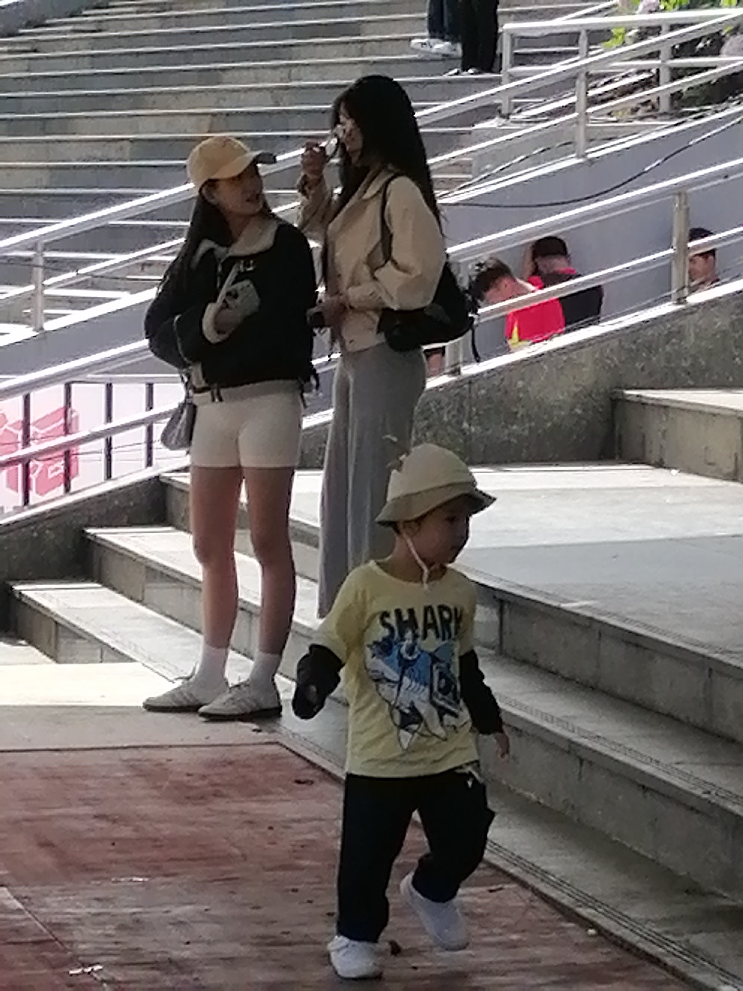 The image depicts a group of people on a tiered concrete structure, possibly a stadium or outdoor seating area, with metal railings. In the foreground, a young child wearing a yellow long-sleeved shirt with a "SHARK" graphic and dark pants, and a wide-brimmed hat, walks down a red-toned walkway. Behind the child and to the left, two young women are standing on the concrete steps. One woman, wearing a light-colored jacket and grey pants, is facing away from the camera. The other woman, on the left, is wearing a black jacket, white shorts, white socks, and white sneakers, and has a light-colored baseball cap on. She is holding a bag and looking towards the woman in the grey pants. In the background, other individuals are visible on the stairs and behind the railings, some of whom appear to be watching or interacting. The overall environment suggests an outdoor public space with bright sunlight casting shadows. The scene does not provide any specific indicators of the city of Da Lat, Vietnam.