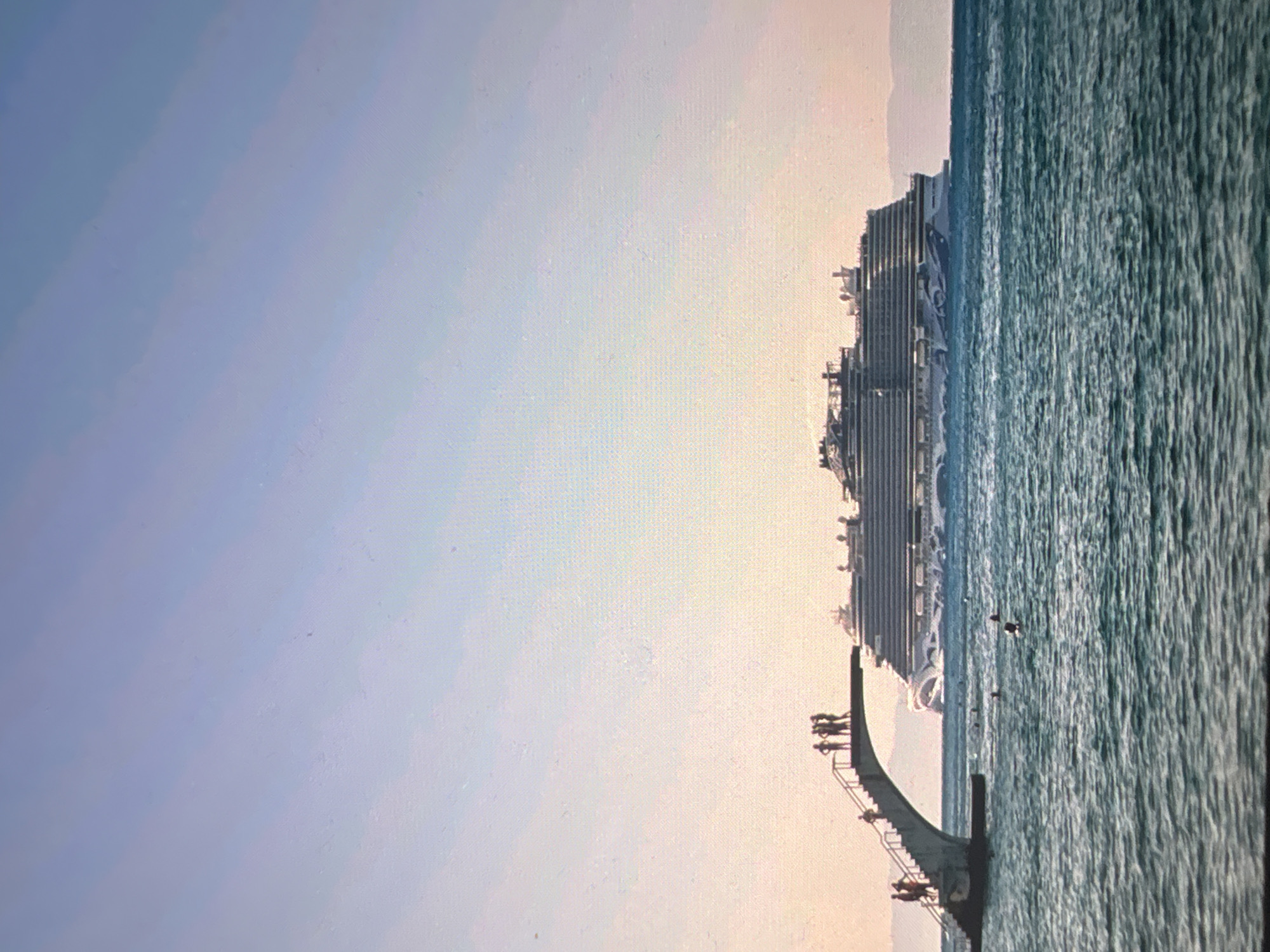 A serene coastal scene captured during what appears to be late afternoon or early morning. A large, multi-deck cruise ship, primarily white with dark accents, dominates the right side of the image, floating on calm, rippled blue-green water. Its silhouette is sharp against the bright horizon.
In the foreground, a gracefully curved pier or jetty extends from the bottom-left of the frame, leading the viewer's eye towards the cruise ship. Several small figures, likely people, are visible along the length of the pier, some appearing to be observing the ship or the surrounding seascape.
The sky, filling a significant portion of the upper-left, displays a gentle gradient, transitioning from a soft, warm peach or light golden hue near the horizon line to a pale, clear blue overhead. This color transition suggests either sunrise or sunset. Distant landmasses or mountains are faintly visible along the horizon behind the ship, adding depth to the scene. The water is calm, showing subtle reflections and textures. There is no visible text in the image. The overall impression is one of tranquility and vastness.
