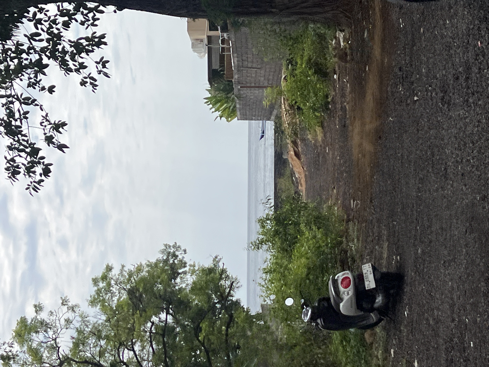 A dirt path leads towards the ocean, bordered by lush green foliage and trees. A black and silver scooter is parked on the left side of the path. In the distance, the calm ocean stretches to the horizon under a cloudy sky. A small blue boat is visible on the water. To the right, a stone wall encloses a residential property with a modern building. The scene appears to be in a coastal area.