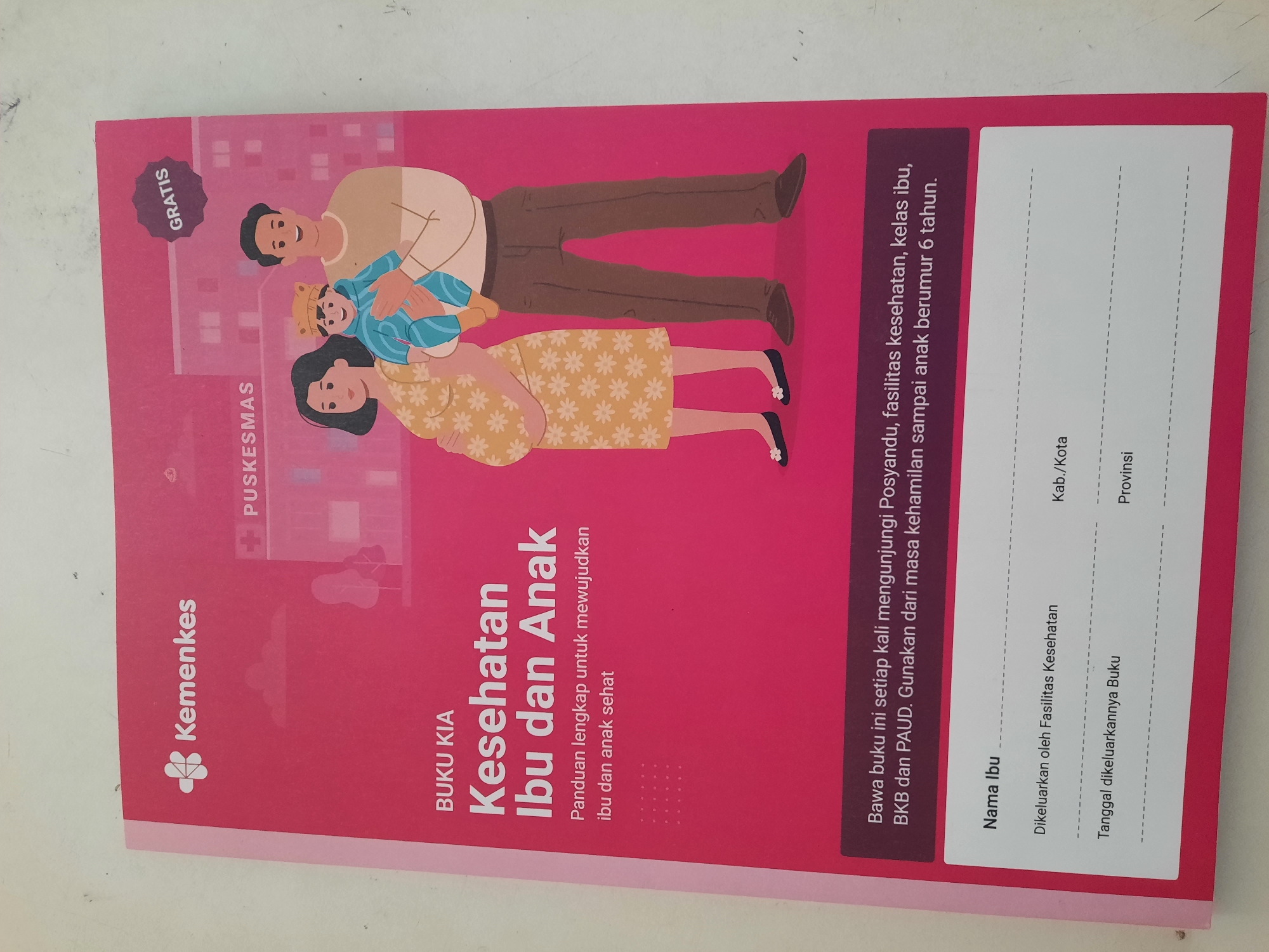 The image shows the cover of a "Buku KIA" (Mother and Child Health Book) from the Indonesian Ministry of Health (Kemenkes). The cover features a graphic of a pregnant woman, a man holding a baby, and a building labeled "PUSKESMAS" (Community Health Center). There is also a "GRATIS" (Free) sticker in the upper right corner. The lower portion of the cover has fields for "Nama Ibu" (Mother's Name), "Dikeluarkan oleh Fasilitas Kesehatan" (Issued by Health Facility), "Tanggal dikeluarkannya Buku" (Date the Book was Issued), and spaces for "Kab./Kota" (Regency/City) and "Provinsi" (Province).
There is no information in the image that specifies a location in the city of Lhokseumawe, Indonesia.