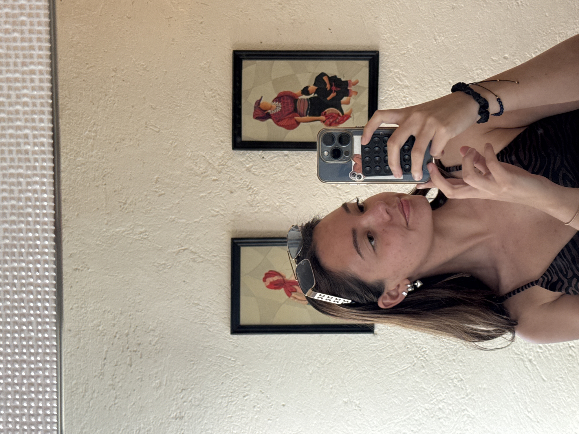 A person is taking a mirror selfie in a room with textured off-white walls. The person is wearing sunglasses in their hair, dangling earrings, and a tank top with a black pattern. They are holding a smartphone with a case, facing the camera. On the wall behind them are two framed pictures of people with bright red clothing and dark hair. A white shutter-style covering appears to the left. The location is not specified in the prompt. The prompt only mentions Puebla, Mexico.