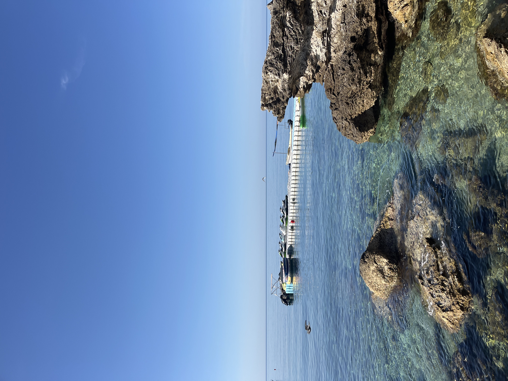 This detailed image captures a serene coastal scene in Budva, Montenegro, under clear daytime conditions. The upper third of the frame consists of a cloudless, uniform light blue sky, with the exception of a faint, wispy, S-shaped white cloud formation located in the upper-right quadrant. A tiny dark speck, possibly a bird or insect, is visible below and to the left of this cloud.
The middle and lower two-thirds of the image are dominated by very clear, calm turquoise waters. The water's transparency allows visibility of the rocky seabed and submerged rock formations in the foreground. The surface exhibits minimal ripples, reflecting the blue sky. A distinct, flat horizon line separates the sky and the sea, positioned approximately one-third of the way down from the top.
The foreground and right side of the image feature prominent, jagged, dark brown and grey rock formations, displaying textured and weathered surfaces. One large rock formation on the right extends into the water, forming an overhang. Submerged rocks are visible through the transparent water in the immediate foreground.
In the mid-distance, a white modular floating dock is anchored horizontally. Moored alongside this dock are several watercraft: at least five personal watercraft (jet skis), primarily white with green accents, and one with yellow and black colors. A small, light-colored motorboat with an outboard engine is also tied to the dock. Adjacent to the motorboat, a partially inflated yellow and blue circular inflatable is visible, bearing the partially visible text "WWW.ADRENALINE.GE". Further behind the main dock, a small green boat or kayak is discernible, and another light-colored boat with a dark, possibly canvas, canopy is visible further to the left.
A single bird, resembling a seagull with light underparts and dark wings, is captured mid-flight in the mid-left portion of the image, above the clear water and in front of the dock.