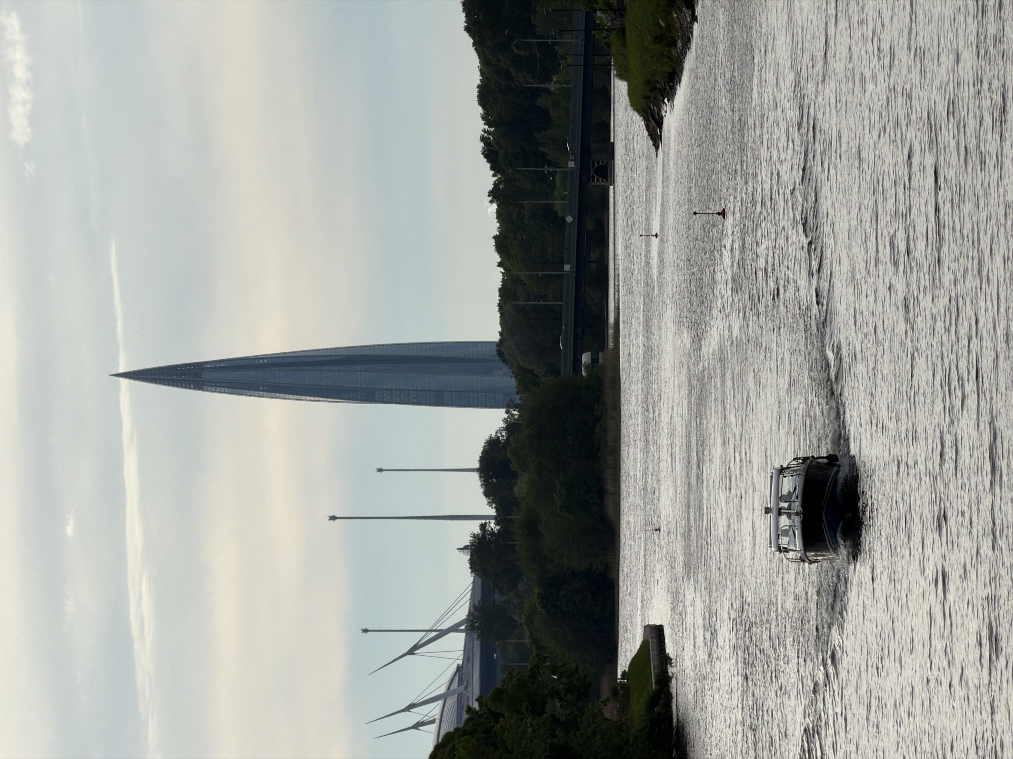 The image displays a modern urban landscape in Saint Petersburg, Russia, centered around a wide waterway, likely a river or canal. The most striking feature is the Lakhta Center, a tall, slender, and pointed skyscraper, dominating the left side of the frame with its sleek glass facade.
On the right side of the waterway, a small, dark motorboat is visible, moving away from the viewer and creating a distinct wake on the textured surface of the water. The water itself shows numerous ripples and reflections, indicating movement and light. Two small, red and white buoys or markers are sparsely placed in the water.
The banks of the waterway are lined with a dense expanse of green trees and foliage. Beyond the treeline on the left, parts of a large cable-stayed bridge, consistent with the Western High-Speed Diameter, can be seen, with its distinctive white pylons rising towards the sky.
The sky above is bright, with soft, diffused light, suggesting either early morning or late afternoon. There are some light clouds and a single, long contrail stretching across the upper left portion of the sky. The overall scene is calm, showcasing a blend of impressive modern architecture, significant infrastructure, and natural elements along the city's waterfront. No visible text or people are discernible.