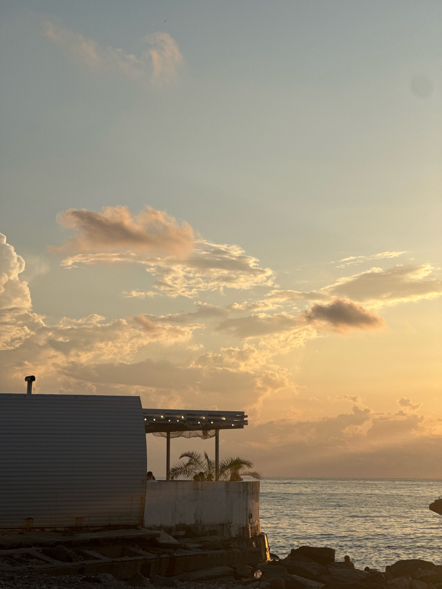 A vertical, low-angle shot captures a coastal scene at dusk in Batumi, Georgia. The sky transitions from a pale blue at the top to a warm orange glow near the horizon, filled with scattered, textured clouds. In the foreground, a low-lying structure with a corrugated metal facade extends from the left side of the frame. Adjacent to it is a concrete platform with a partially visible covered seating area adorned with string lights. Two palm trees are planted behind a low concrete barrier, and a few silhouetted figures are visible in the seating area. The sea, a deep blue with gentle waves, occupies the right side of the image, meeting a rocky shoreline in the lower right corner where more faint silhouettes of people can be discerned. The overall impression is one of tranquil evening by the sea.