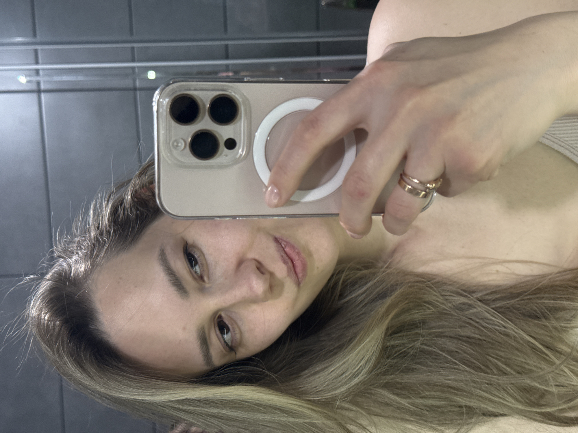 A woman with light brown hair is taking a selfie in a bathroom. She is holding a gold iPhone in her right hand, which has a clear case and a white MagSafe ring on the back. She is wearing two rings on her ring finger. The background shows gray tiled walls. The location cannot be determined as Chaikovsky, Russia based on the provided image.
