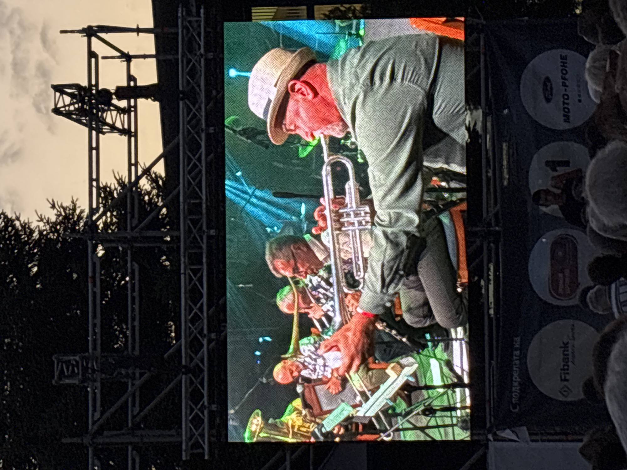 The image shows a large outdoor screen displaying a live performance by a band. The band members are playing various instruments, including trumpets and percussion. In the foreground, a man in a green shirt and a straw hat is playing a trumpet. The background features stage lighting in blue and green hues. Below the screen, there are banners with logos, including "Fibank" and "MOTO-PFOHЕ". The overall setting appears to be a festival or concert. The presence of the logos and the setup suggest an event organized by sponsors, possibly at an outdoor venue. The event likely took place during the evening or late afternoon, given the lighting conditions. The details on the banners and the context of a music performance suggest this is an event in Bansko, Bulgaria, likely part of a cultural festival or a dedicated music event.