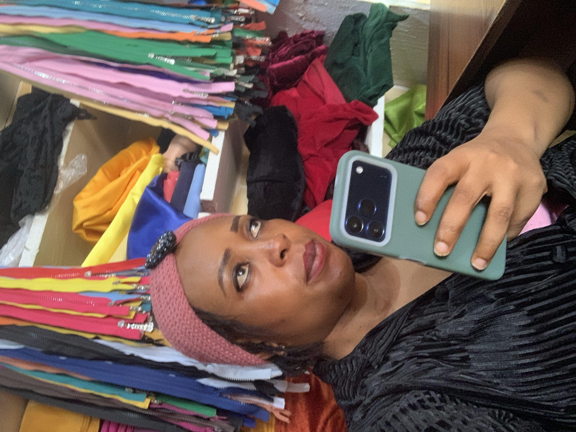 A woman, identified as residing in Yelwa, Nigeria, is pictured holding a green smartphone in her right hand, which is positioned in the lower right quadrant of the frame. She is looking towards the right, with her gaze slightly upward and her mouth slightly open, suggesting she might be speaking or listening. She wears a black, long-sleeved garment with a textured, ribbed pattern. Her head is adorned with a pink headscarf, accented with a dark, possibly beaded, embellishment on the left side. The background appears to be a retail setting, likely a fabric or tailoring shop, with shelves stocked with various colorful zippers and folded fabrics in shades of red, green, black, and yellow. The lighting is moderate, casting soft shadows and illuminating the subjects and their surroundings.