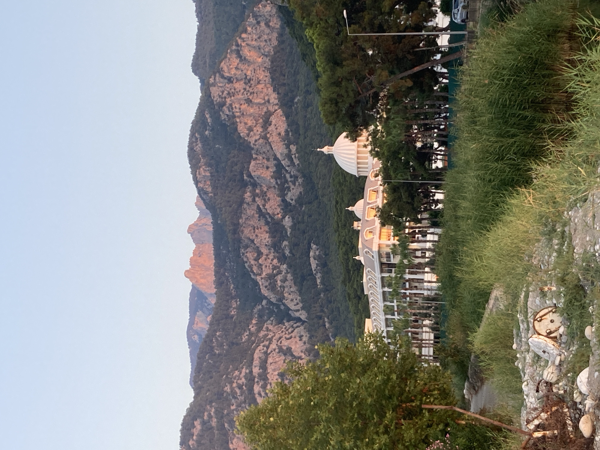 In Kemer, Turkey, a resort hotel with white facades and a prominent white dome stands amidst lush greenery, including palm trees and tall reeds. The hotel is backed by a dense forest that extends up the slopes of a mountainous landscape. The mountains themselves are rugged with exposed rock faces, some of which are illuminated by the warm glow of sunset, creating a striking contrast with the shadows cast by the trees. In the foreground, a grassy embankment leads down to a small clearing strewn with debris, including discarded metal objects and stones. The sky above is a clear, pale blue, indicative of early evening.