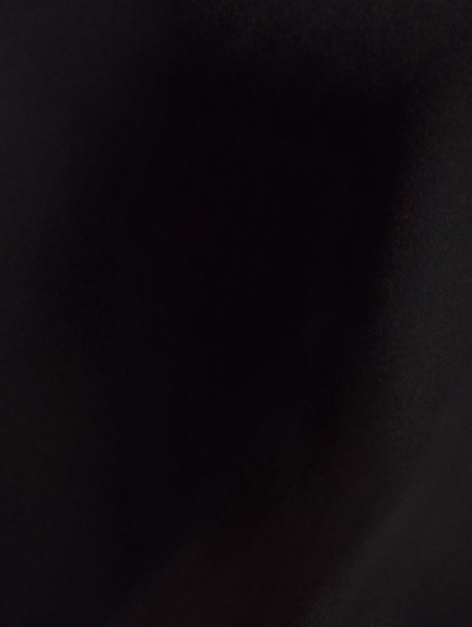 The image is a close-up, dark, and grainy shot of a black surface. The texture appears slightly rough, with subtle variations in light reflection that suggest it might be fabric, a matte painted surface, or a similar material. There are no discernible objects, people, or environmental features. The lighting is dim, with a brighter area concentrated on the right side of the frame, creating a soft gradient. The location is unknown.