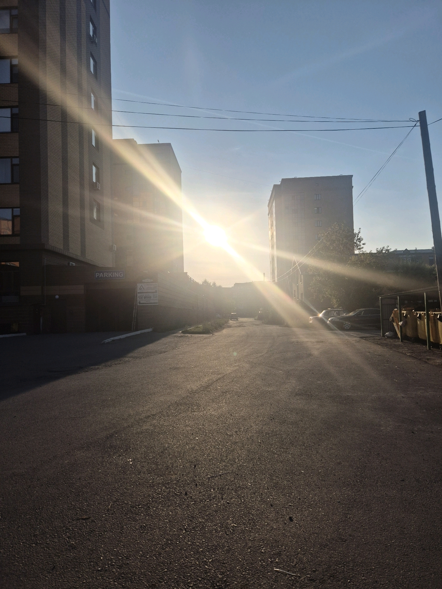 The image depicts a sun-drenched street in Kokshetau, Kazakhstan, likely in the early morning or late afternoon given the low angle of the sun and the long shadows cast. Two tall, multi-story buildings dominate the scene, one on the left and another further down the street on the right. The building on the left features a brick facade and windows, with a sign indicating "PARKING" above an entrance. The building on the right appears to be of similar construction, also with multiple windows.
The asphalt road stretches towards the horizon, with a distinct shadow line dividing the sunlit portion from the shaded area. Several cars are parked along the right side of the road, partially obscured by sunlight and shadows. A row of yellow bins or containers is visible near the parked cars. Overhead, power lines are strung across the sky, supported by a utility pole on the far right. The sky is a clear blue with a few faint wisps of clouds. The overall impression is one of a quiet urban environment bathed in warm, golden light. There are no people visible in the image.