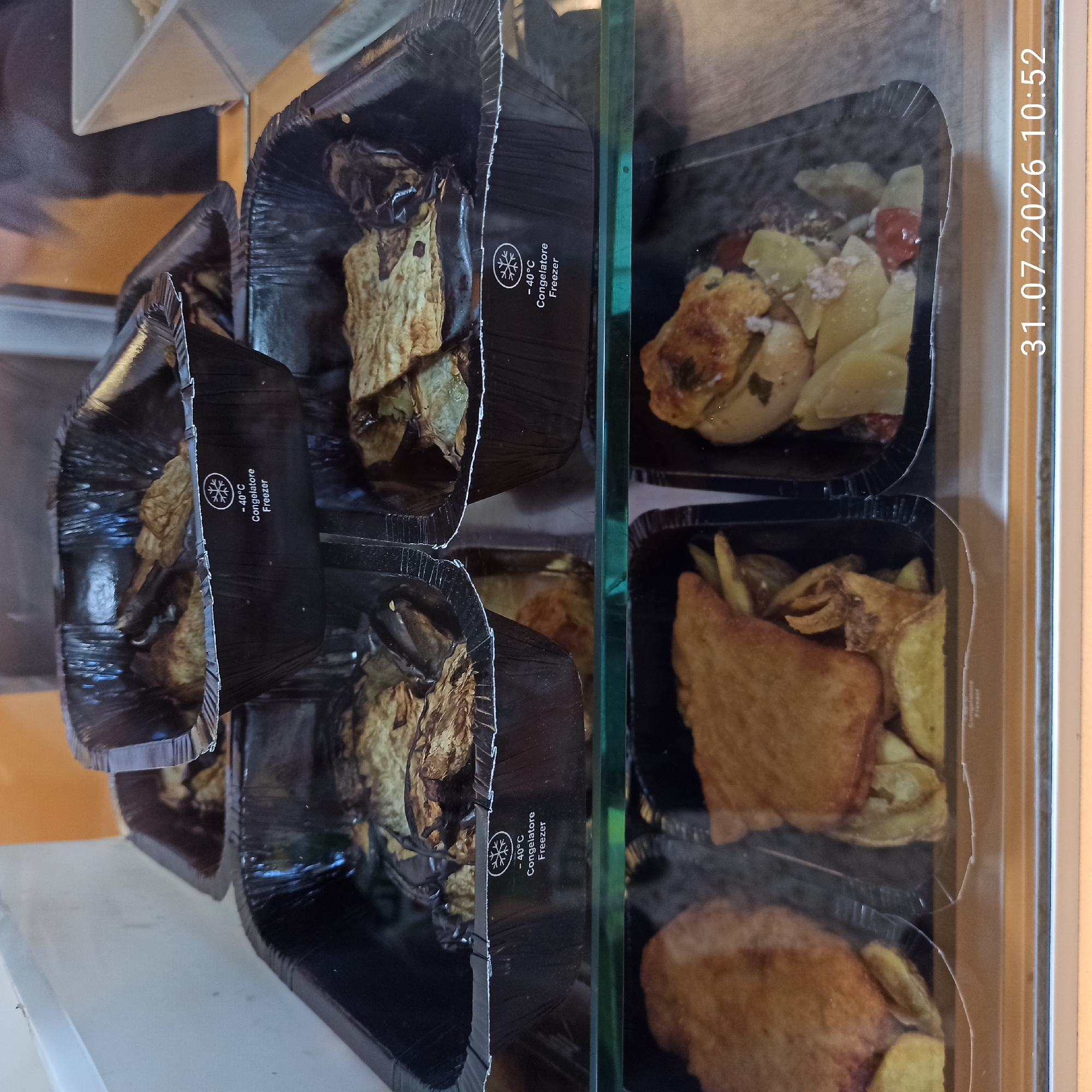 The image displays a close-up view of several food containers, likely from a deli or prepared food section, arranged on shelves. The containers are black and appear to be made of foil or a similar material. The top containers hold what seems to be fried or baked eggplant. Below these, two containers are visible, one containing what looks like fried chicken or schnitzel with fries, and the other containing a stew-like dish with vegetables and possibly meat. Some of the containers have labels indicating "-40°C Congelatore Freezer". The overall setting appears to be a refrigerated display case, suggesting a commercial food establishment. A timestamp "31.07.2026 10:52" is visible in the bottom right corner of the image. There is no information in the image that can determine the location in the city of Modugno, Italy.