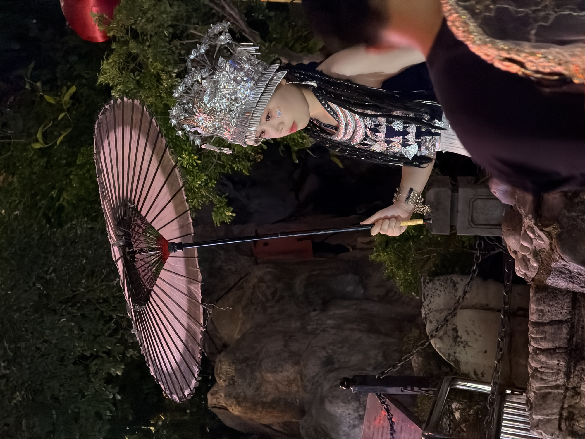 A young East Asian female wearing a decorative silver headdress, elaborate necklaces, and dark clothing is holding a pink umbrella. She has dark braided hair and glitter makeup on her face. She is standing in front of a rocky embankment and lush green foliage. Behind her, a large red spherical object is visible amongst the trees. To the right, the back of another person's head is visible, also wearing similar decorative attire around their neck. The scene appears to be outdoors at night, possibly in a park or outdoor event space, with artificial lighting illuminating the subjects and surroundings. The location is identified as Yuzhong District, China.