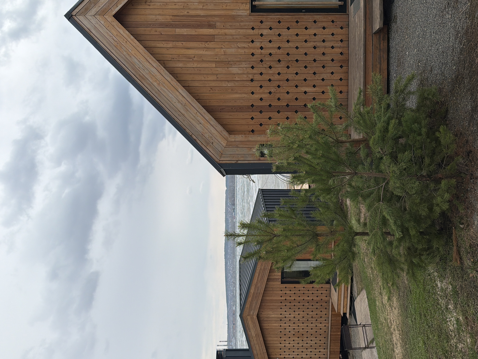 The image depicts a scene in Votkinsk, Russia, featuring two modern wooden buildings with gabled roofs. The buildings exhibit decorative diamond-shaped cutouts in their wooden facades. Between the buildings, a body of water, possibly a river or lake, is visible, with some industrial structures along the shoreline. Overcast skies are present. In the foreground, a small coniferous tree partially obscures one of the buildings. The ground appears to be covered with grass and some gravel or mulch. The composition is taken from a slightly elevated viewpoint.