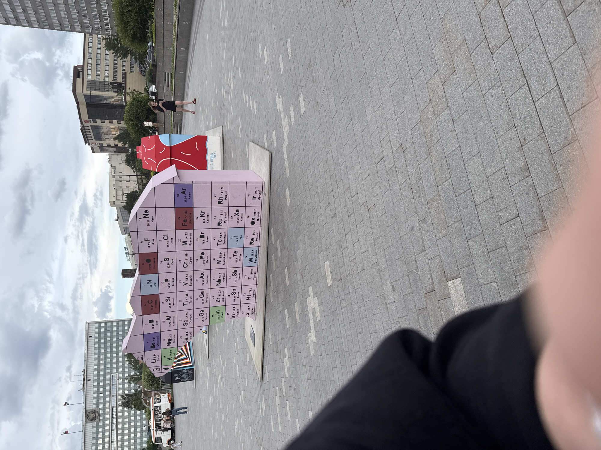 This is a photograph taken outdoors in a city square, likely on a cloudy day. The main subject is a large, colorful, three-dimensional sculpture resembling a stylized periodic table of elements. It's pink and has elements depicted on its surfaces with their symbols, atomic numbers, and names in Russian. Some sections are red and blue. Next to it, there's another abstract sculpture in red and blue.
In the background, there are modern buildings and trees. A few people are visible in the distance, including a woman in a black dress walking near the sculptures and a group of people near a white van in front of one of the buildings.
The image is oriented vertically. The perspective suggests the photo was taken from a low angle, possibly by someone holding a phone or camera. Part of the photographer's hand and arm, wearing a black garment, is in the lower right corner, obscuring a portion of the scene. The ground is paved with grey square tiles. Some text is visible on the red and blue sculpture: "ПЕРНОД 2025".
