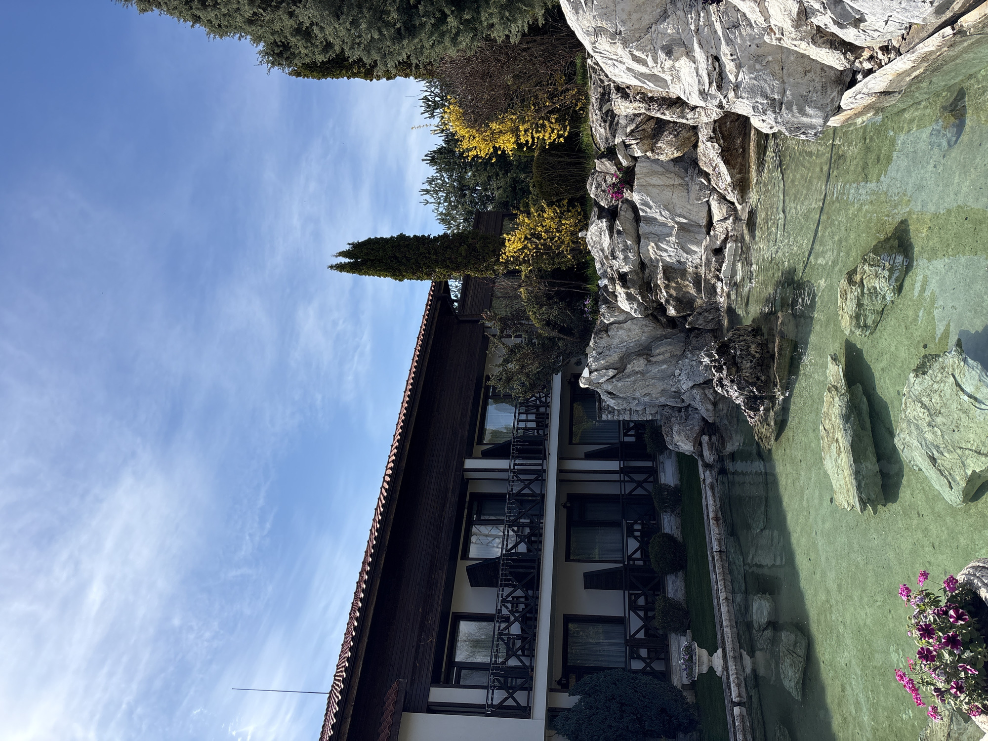The image depicts a building complex near a body of water, possibly a pond, in Koprivlen, Bulgaria. The building has a brown roof, a white facade, and several windows with balconies. Trees and rocks surround the pond. The sky is blue with scattered clouds. The photograph appears to be taken during the daytime. There are flowering plants visible near the water.