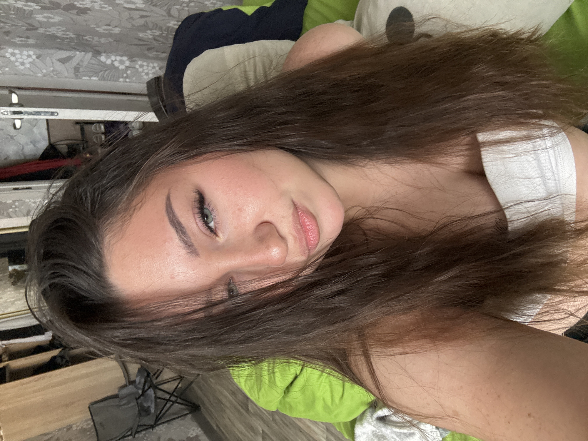 A young woman with long, dark brown hair, green eyes, and light skin is lying down on a bed or couch with a bright green cushion. She is wearing a white bandeau top. Her hair partially covers her face. In the background, there is a closet with clothing hanging, a folding chair, and patterned wallpaper. The lighting appears to be indoor. There is no indication of the location being in the city of Perm, Russia.