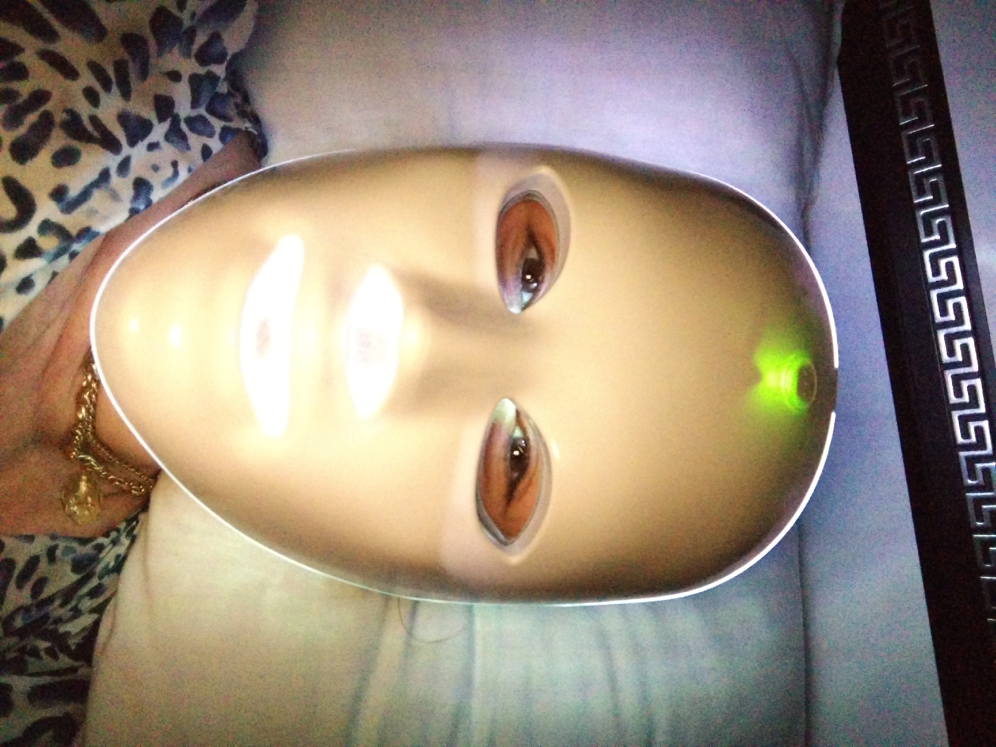 The image captures a close-up, eye-level perspective of an individual lying down and undergoing a facial treatment. The central subject is a person wearing a smooth, light-toned, full-face LED light therapy mask. The mask features oval-shaped cutouts through which the person's dark brown eyes are visible. A prominent, bright green light illuminates the forehead area of the mask, while a bright white light emanates from the mouth area. The periphery of the mask exhibits a subtle white glow.
The person is wearing a garment with a dark and light, possibly animal-print, pattern visible on the chest and shoulder. A golden-colored chain necklace with a small, circular or spherical pendant is visible around their neck.
The background above the person features a dark, horizontal object, likely a headboard or furniture element, adorned with a repetitive, metallic Greek key or meander pattern. The immediate environment around the person's head appears to be a light-colored pillow or bedding. The overall lighting of the scene is dim, with the mask's illumination being a primary light source. The distant background exhibits muted tones and a slight graininess, consistent with low ambient light conditions.