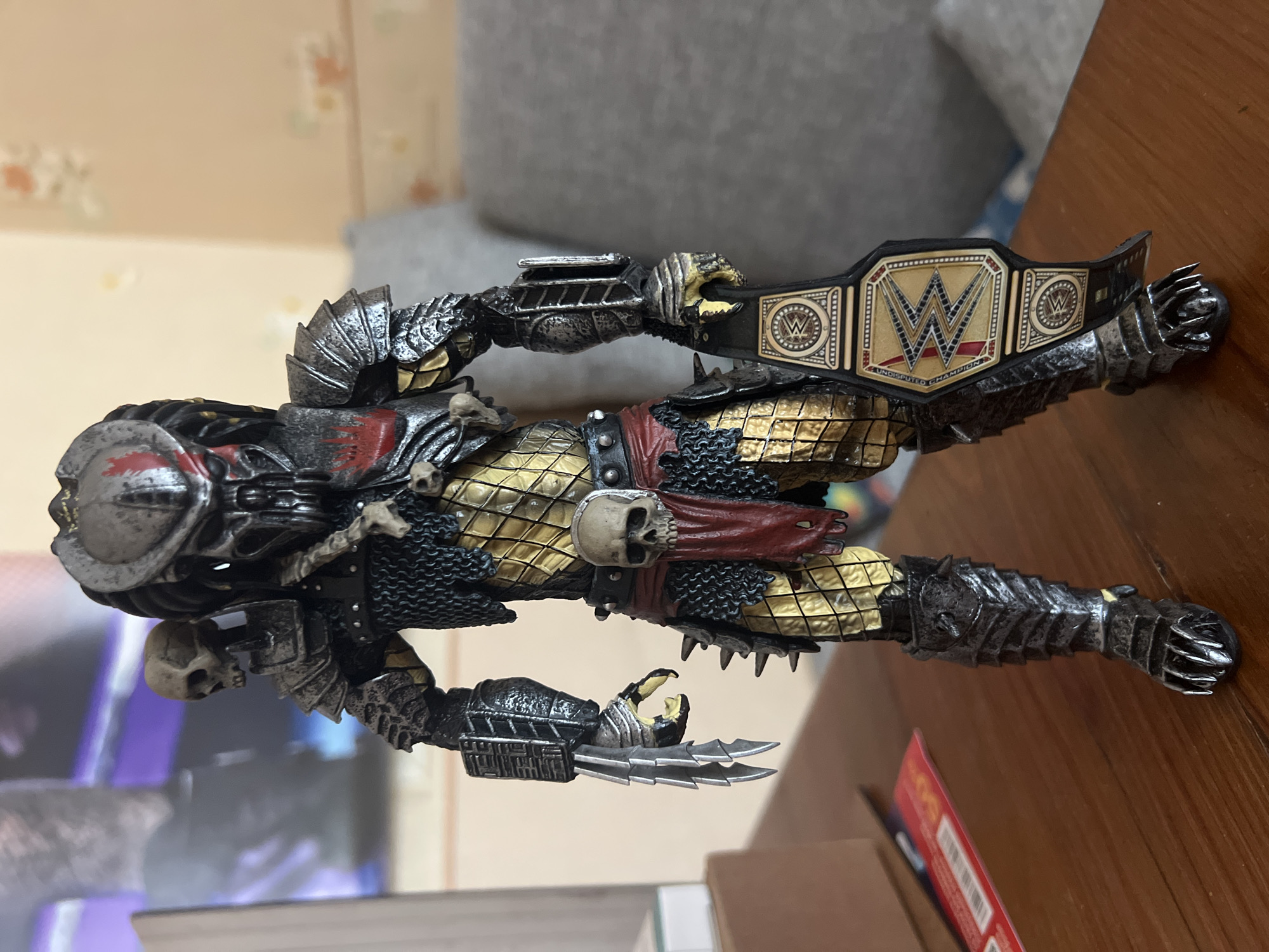 A close-up, eye-level shot shows a Predator action figure standing on a wooden table. The figure is detailed with armor, tribal markings, and a skull accessory on its shoulder. It holds a WWE championship belt in its right hand. The background is blurred, showing a wall with an abstract pattern and a couch. The lighting is even, and the focus is sharp on the action figure. The scene is indoors, likely in a home setting, given the presence of furniture and wall decor. The wooden table surface is smooth and appears to be made of polished wood. To the left of the figure, a partially visible object with a white and red label rests on the table.