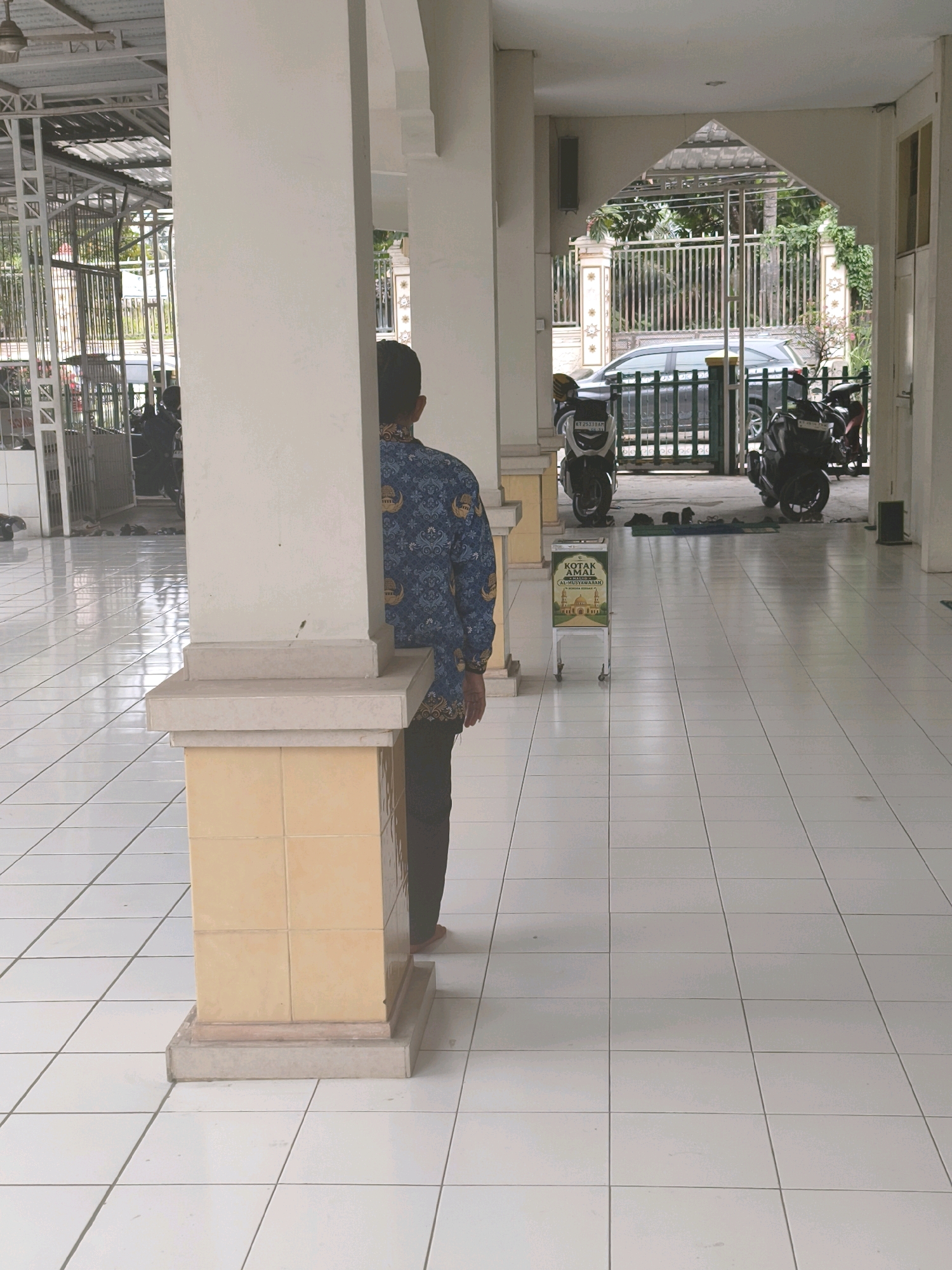The image captures a covered walkway with white tiled flooring, leading to an open area with a gate. In the foreground, a person is partially visible, standing behind a pillar. They are wearing a dark blue patterned shirt and dark pants. To the left, a portion of a building structure with a metal roof and fencing is visible. In the background, through the gate, a car and several motorcycles are parked. A donation box with text in Indonesian is positioned in the walkway. The environment suggests a public space, possibly a mosque or community center, in Samarinda, Indonesia, given the architectural style and the presence of Islamic script on the donation box.