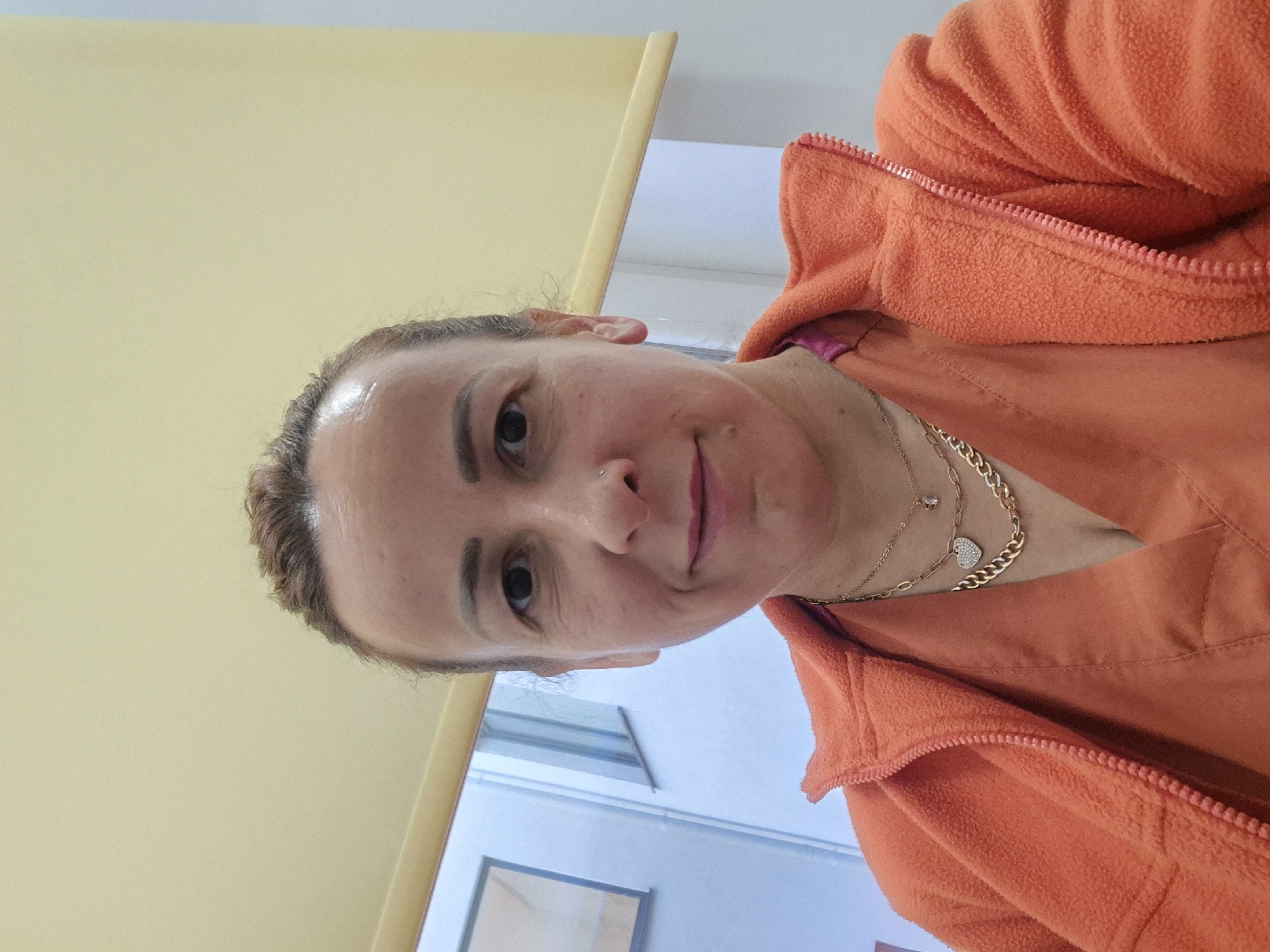 Here's a detailed description of the image:
**People:**
* A woman with fair skin is the primary subject. She has brown hair pulled back, and visible fine lines around her eyes. She is wearing a gold necklace. She has a piercing on her nose.
**Objects:**
* She is wearing an orange fleece zip-up jacket.
* A cream-colored wall is visible in the background
* A window with a light blue frame is visible in the background
**Environment:**
* The setting appears to be indoors, possibly a medical facility or office. The city location is unknown.
**Actions and Interactions:**
* The woman appears to be taking a selfie.
**Overall Impression:**
The image has a casual, informal feel, likely taken in a private setting.