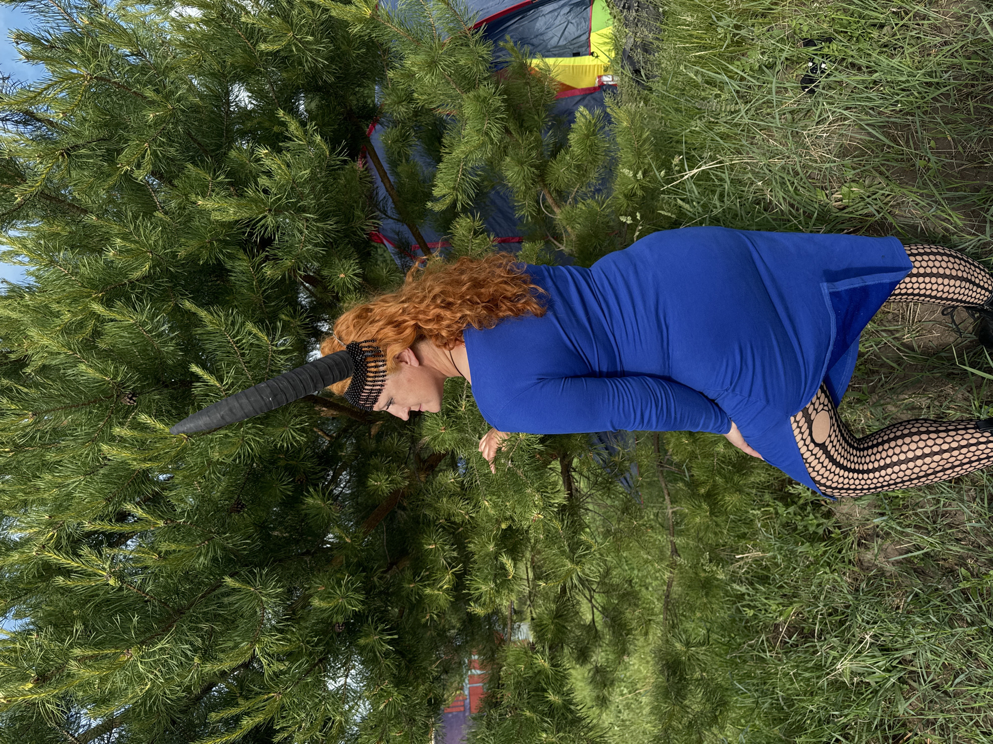 The image displays a woman lying on her side in an outdoor setting, positioned alongside a large pine tree and grassy ground. She has vibrant curly red hair and wears a long-sleeved royal blue dress. A distinctive black horn-like headpiece, adorned with a beaded band, rests on her head. Her legs are clad in black fishnet stockings featuring a polka-dot pattern, with dark footwear partially visible.
A dense pine tree with green needles occupies a significant portion of the left and central frame. On the right, green grass covers the ground. In the background, partially obscured by the tree, a colorful tent with blue, yellow, red, and green sections can be seen, suggesting a camping or outdoor recreational area. A clear blue sky is visible in the upper left corner.
The scene takes place during the daytime, under bright natural light, indicating sunny weather. The woman appears to be posing calmly. There is no visible text in the image. The setting suggests a natural environment, likely in Большая Кивара, Russia.