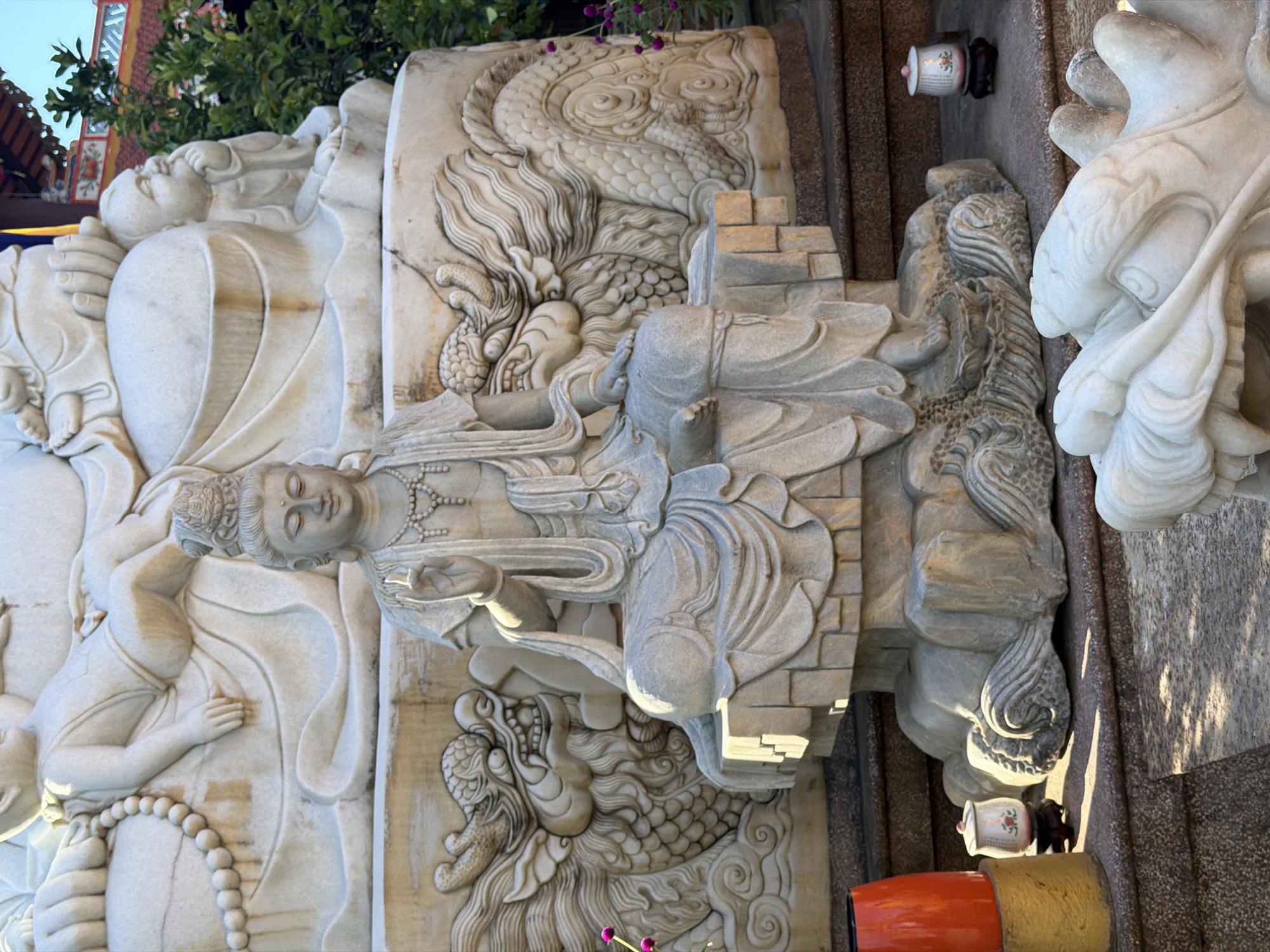 The image captures an intricate stone carving in Đà Nẵng, Vietnam. The carving depicts a reclining female figure adorned with jewelry, possibly a deity or a religious figure. The figure rests on an architectural structure, surrounded by detailed depictions of dragons. Additional figures are carved into the stone on either side. To the right of the carving, there are ornamental objects: a small ceramic container with floral design and an orange and gold cylindrical structure. The backdrop includes fragments of buildings and greenery, suggesting an outdoor temple or garden setting. The carving's weathered appearance indicates age and exposure to the elements.