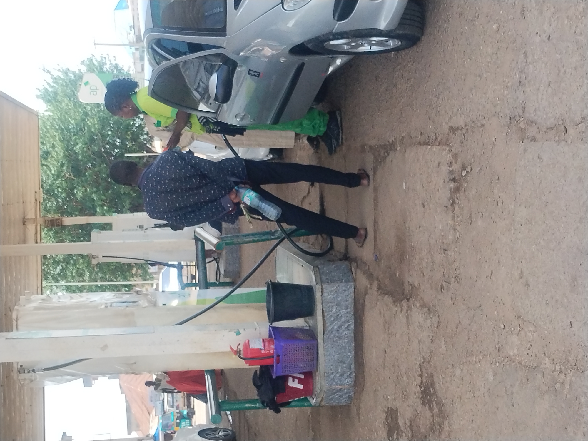 The image shows a scene at what appears to be a gas station in Jos, Nigeria. Two individuals are present. One, a person with dark skin and braided hair, is wearing a bright lime-green shirt and dark pants. They are standing near the open driver's side door of a silver car, which is partially obscured. The other individual, a man with dark skin, is wearing a dark patterned shirt and dark pants. He is holding a transparent plastic bottle filled with liquid.
The scene is outdoors, with a rough, dirt-like ground surface. There are structures resembling part of a canopy or building, with white vertical posts and horizontal beams. In the background, green trees are visible. Several objects are placed on a low, stone-like structure. These include a black bucket, a purple basket, and a red fire extinguisher. Another red object, possibly a bag or fabric, is visible on the ground.
The overall lighting suggests it is daytime, possibly mid-morning or afternoon. The weather appears to be fair. The presence of the car, gas pump hoses (though not clearly depicted in use), and the fire extinguisher suggest a functional gas station. The interaction between the two individuals and the presence of the car indicate a refueling or service stop.