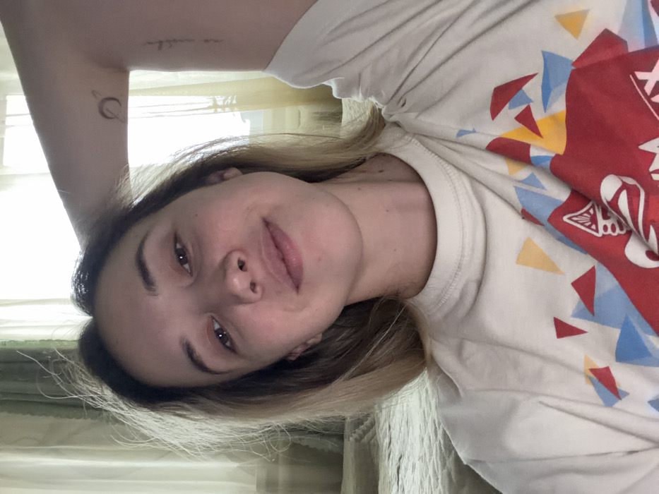 A young adult female is depicted in a selfie taken indoors in Komsomolskiy, Russia. She has light brown hair, dark eyes, and a slight smile, looking directly into the camera. Her left arm is raised, revealing a small tattoo of a ringed planet on her bicep, with a line of text tattooed vertically beneath it. She wears a light-colored, crew-neck t-shirt adorned with a multi-colored geometric pattern of red, blue, and yellow triangles, and a distinct red emblem resembling a stylized cannabis leaf. The background shows a window with sheer light-colored curtains, admitting natural light. To her right, a portion of a bed or sofa with a light-colored, quilted blanket is visible.