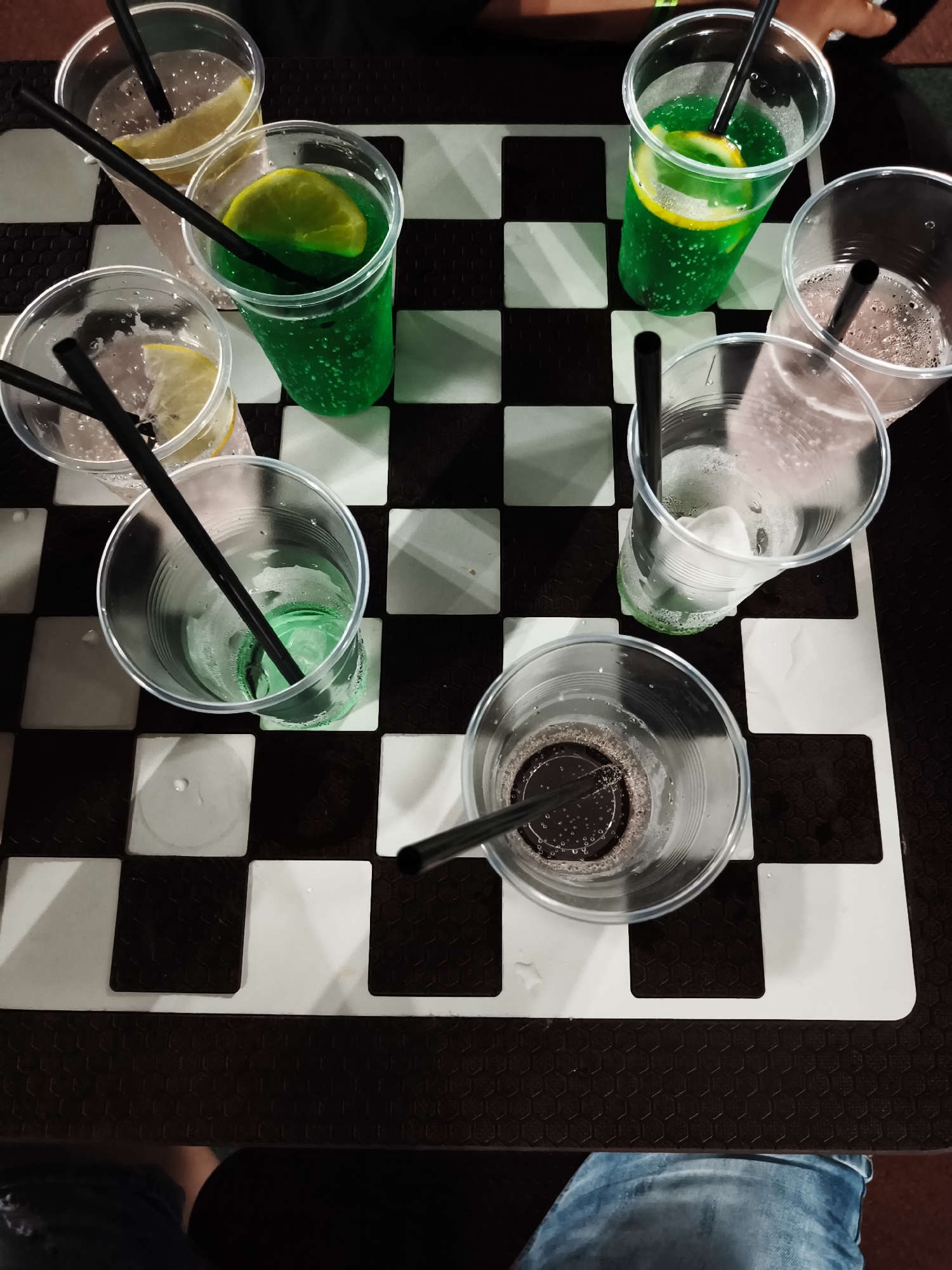 An overhead, high-angle medium shot captures seven plastic cups filled with various beverages and black straws. The cups are arranged on a black and white checkered surface, resembling a chessboard. One cup contains a vibrant green liquid with lemon slices and bubbles. Another cup holds a clear liquid with a slice of lemon and bubbles. Several other cups contain clear or slightly colored liquids with ice and bubbles. One cup in the foreground contains a dark brown or black liquid. The scene appears to be indoors, possibly at a bar or cafe in Kiten, Bulgaria, given the style of the drinks and presentation. No people are clearly visible in the frame, but a person's legs in blue jeans are visible at the bottom edge of the image.