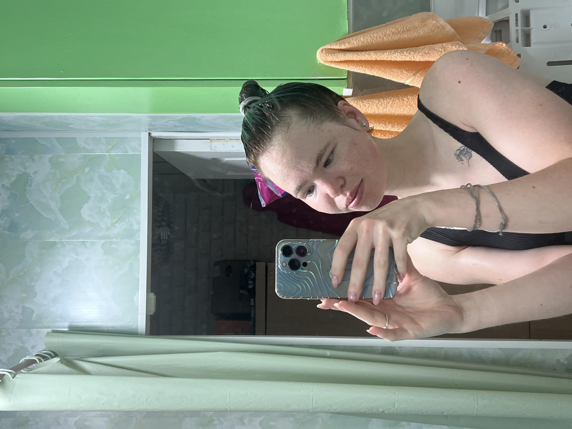 A young woman is captured taking a mirror selfie in what appears to be a bathroom in Chaikovsky, Russia. She holds a smartphone, likely an iPhone Pro model with a distinctive silver and blue wave-patterned case, horizontally in both hands, gazing at the screen. Her dark hair is wet and tied in a bun, showing a noticeable green tint, suggesting recent hair coloring or washing. She has fair skin with some freckles and a small, dark, stylized tattoo (possibly a wave) on her left shoulder/collarbone area. She is wearing a black top or swimsuit, and several silver bracelets are visible on her left wrist, along with a silver ring on her left ring finger. Her fingernails are manicured with light pink polish.
The bathroom features a bright green wall at the top of the frame, with light green and white marbled tiles below it. To the right of the woman, a stack of folded orange towels rests on a shelf. A light green shower curtain or similar fabric is visible at the bottom. The reflection in the mirror shows a darker, less detailed background. The lighting is artificial, characteristic of an indoor setting. There is no visible text in the image.
