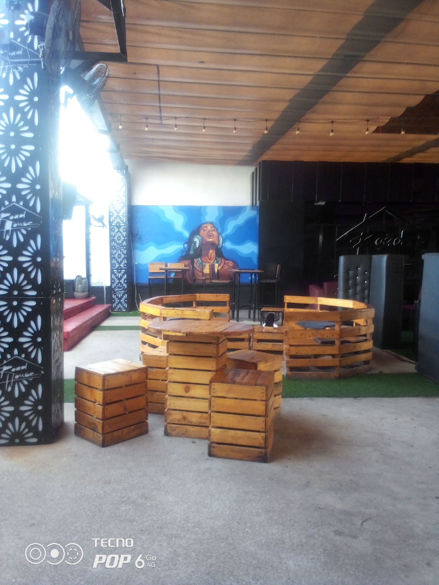 The image depicts an outdoor seating area with rustic wooden furniture, likely a restaurant or bar, situated in Howayi, Nigeria. The furniture is made of stacked wooden crates, forming tables and seating. In the background, a vibrant mural of a woman with headphones adorns a wall, with high-top tables and stools arranged in front of it. The ceiling is constructed of wooden planks, with simple light fixtures hanging from it. On the left side of the image, a decorative pillar with a floral cut-out design and the text "Food House" is visible, next to red-carpeted steps leading upwards. The ground is a concrete surface, with patches of artificial grass visible behind the seating arrangements. The overall ambiance suggests a casual and artistic establishment.